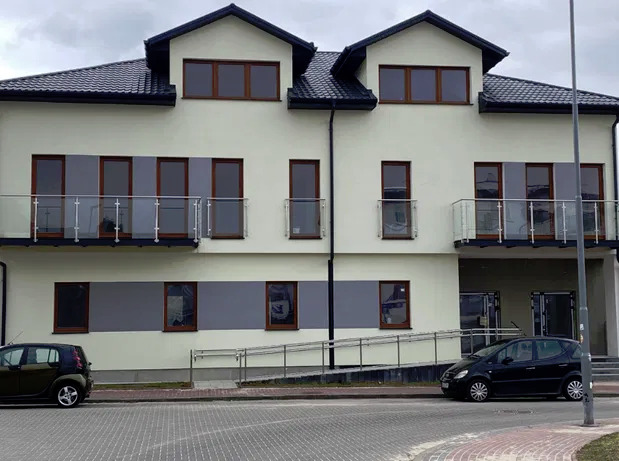 Dwupiętrowy budynek po remoncie z szarą elewacją, brązowymi oknami i czarnym dachem, widoczny z zewnątrz, z zaparkowanymi samochodami.