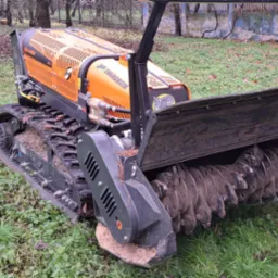 Pomarańczowa, gąsienicowa frezarka do pni w Sieradzu, z widocznymi śladami ziemi na ostrzach i gąsienicach, stojąca na trawniku.