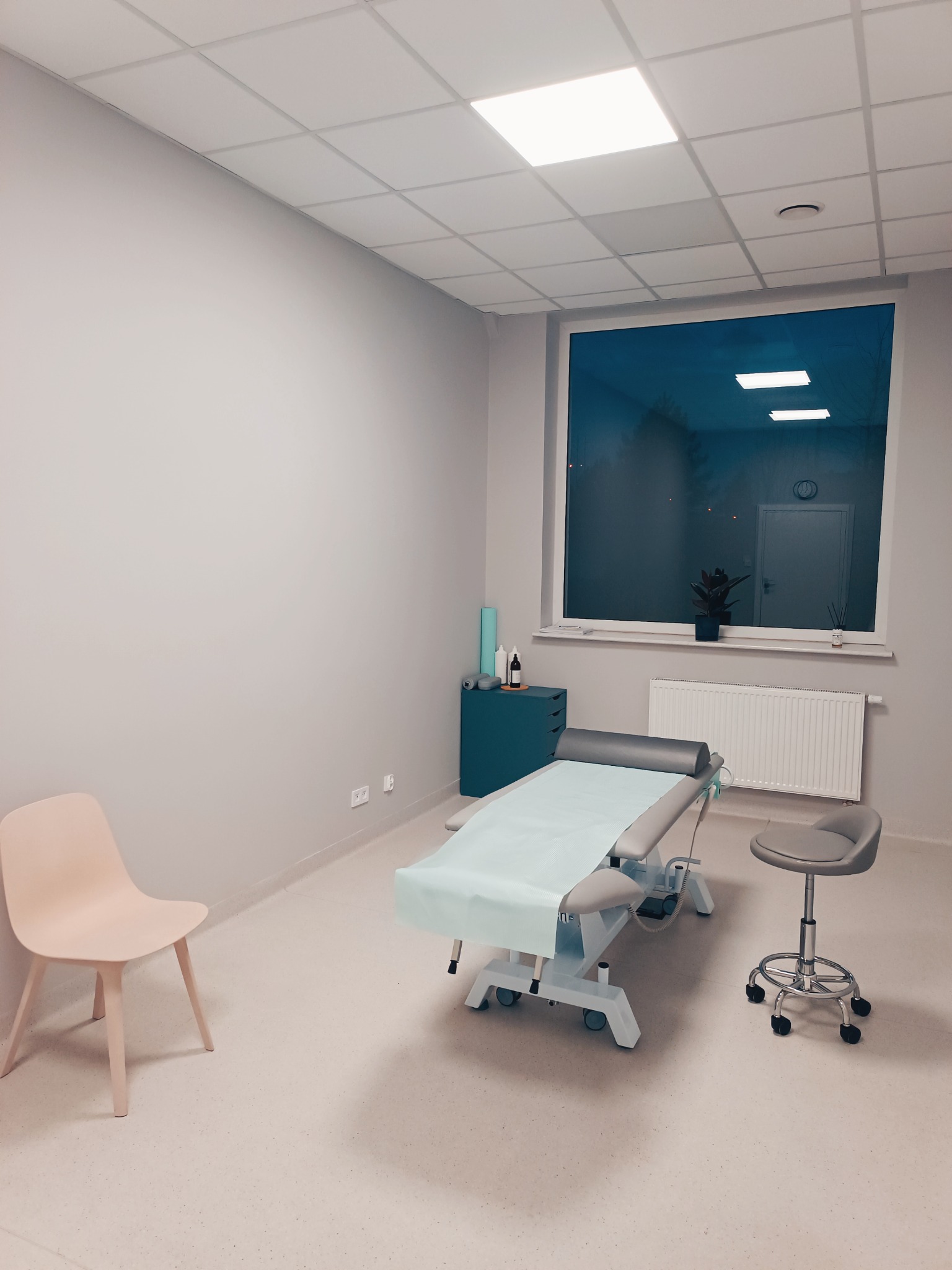 Jasne, minimalistyczne wnętrze gabinetu z kozetką pokrytą jednorazowym prześcieradłem, krzesłem na kółkach i pastelowym krzesłem bez oparcia, widok na okno z rośliną w tle.