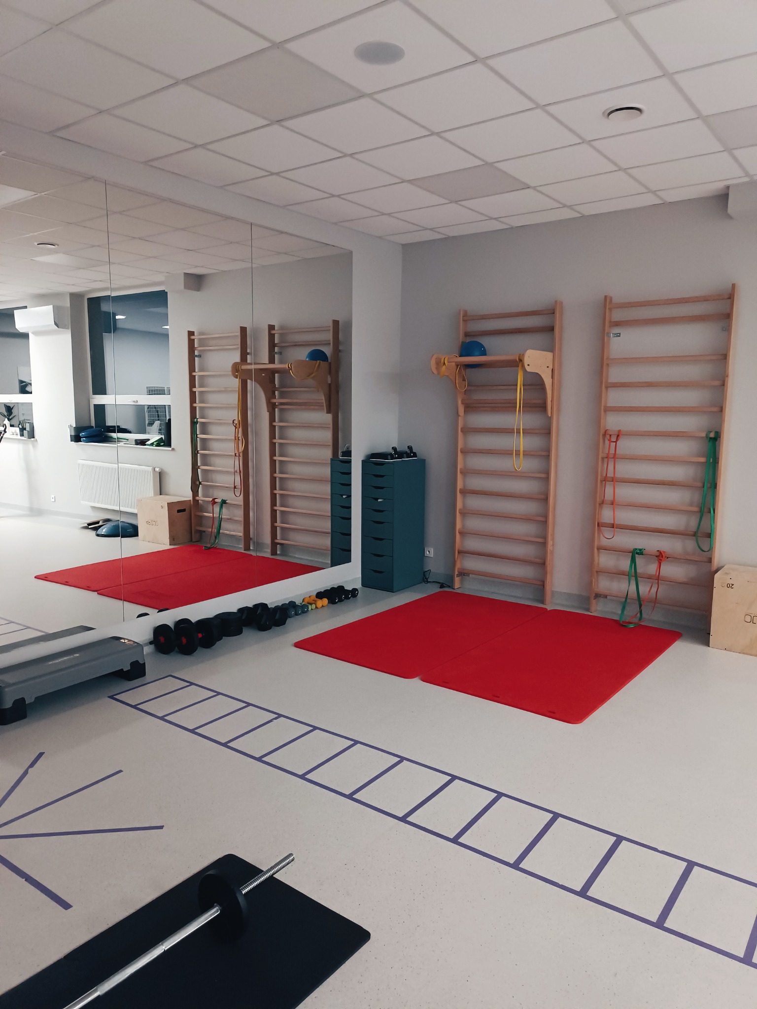 Sala rehabilitacyjna z drabinkami gimnastycznymi, czerwonymi matami, hantlami i stepem, widok z perspektywy podłogi z namalowanymi wzorami.