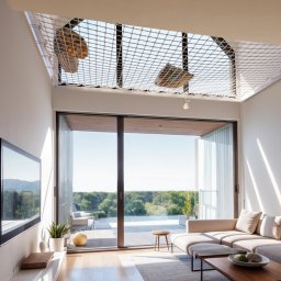 M11 architekci - Jasne wnętrze salonu z siatką relaksacyjną pod sufitem, widok na zewnątrz przez przesuwne drzwi tarasowe, minimalistyczny wystrój z szarą sofą i telewizorem na ścianie.