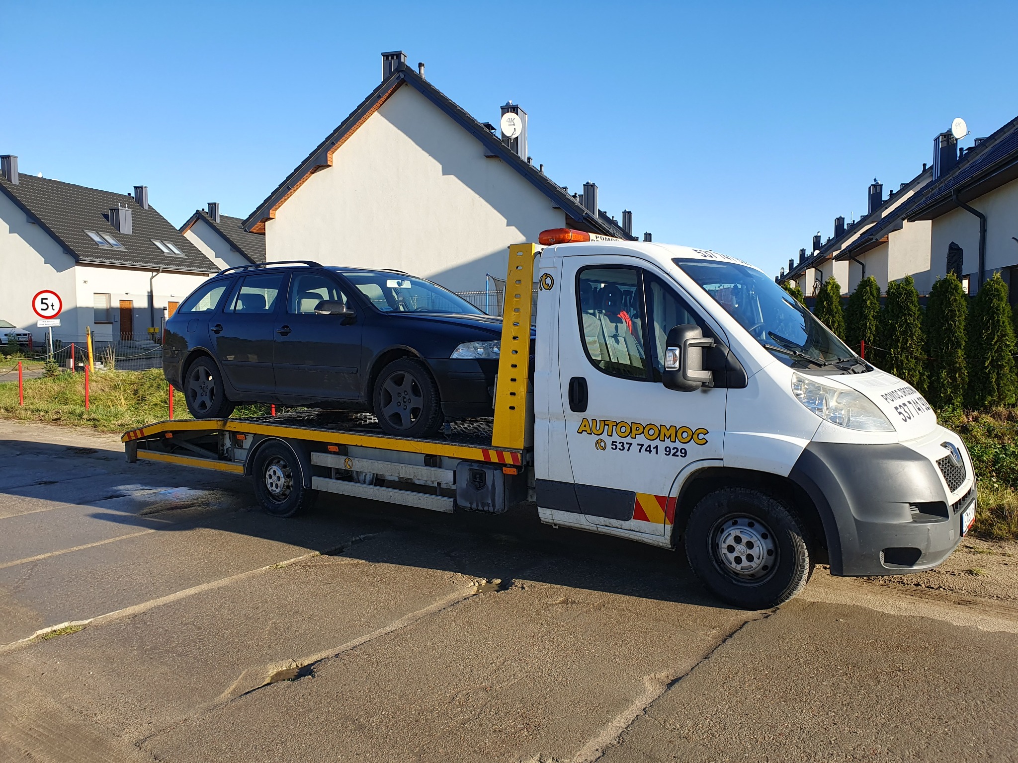 Czarny samochód osobowy na żółtej lawecie autopomocy z numerem telefonu 537 741 929, zaparkowanej na ulicy osiedlowej z rzędem domów w tle.