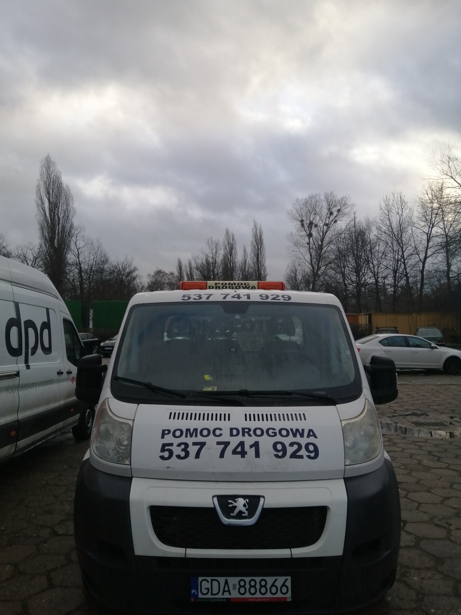 Biały bus pomocy drogowej Peugeot z numerem telefonu i pomarańczowym światłem ostrzegawczym na dachu, zaparkowany na parkingu z kostki brukowej w pochmurny dzień, obok busa DPD.
