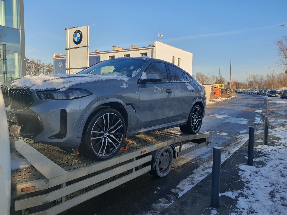Szary samochód BMW X6, częściowo pokryty śniegiem, umieszczony na platformie lawety. W tle widoczny salon samochodowy BMW z charakterystycznym logo.
