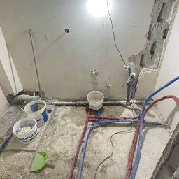 Medallion - Łazienka w trakcie remontu: widoczne rury wodne, wiadro z wodą, pęknięty tynk, tymczasowe oświetlenie budowlane.