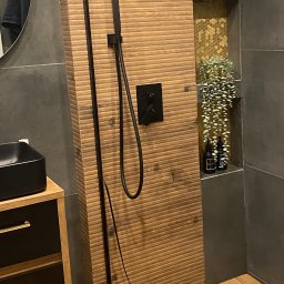 Medallion - Nowoczesna kabina prysznicowa z drewnopodobnymi płytkami na ścianie i podłodze, czarna armatura i panel sterujący, dekoracyjna półka z rośliną zwisającą i kosmetykami.