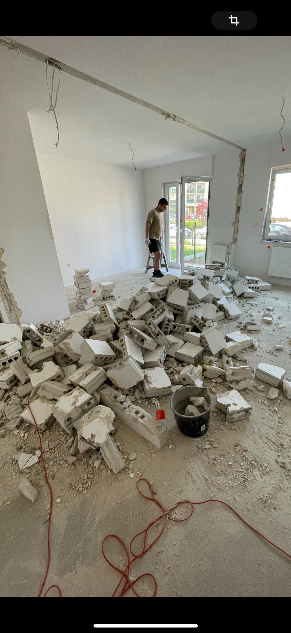 Demontaż ściany z białej cegły w mieszkaniu, widoczne porozrzucane fragmenty, pracownik na drabinie w tle, przewody elektryczne zwisające z sufitu.