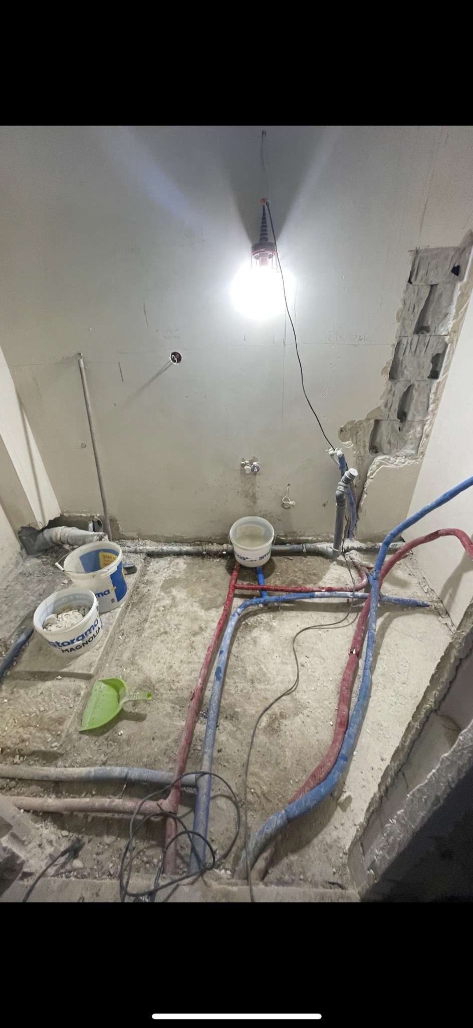 Łazienka w trakcie remontu: widoczne rury wodne, wiadro z wodą, pęknięty tynk, tymczasowe oświetlenie budowlane.