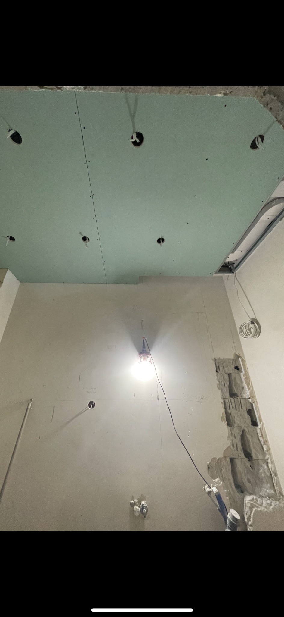 Wnętrze w trakcie prac: sufit z płyt kartonowo-gipsowych z wyciętymi otworami na oświetlenie, fragment ściany z widocznym uszkodzeniem i odsłoniętą instalacją hydrauliczną, oświetlony lampą budowlaną.