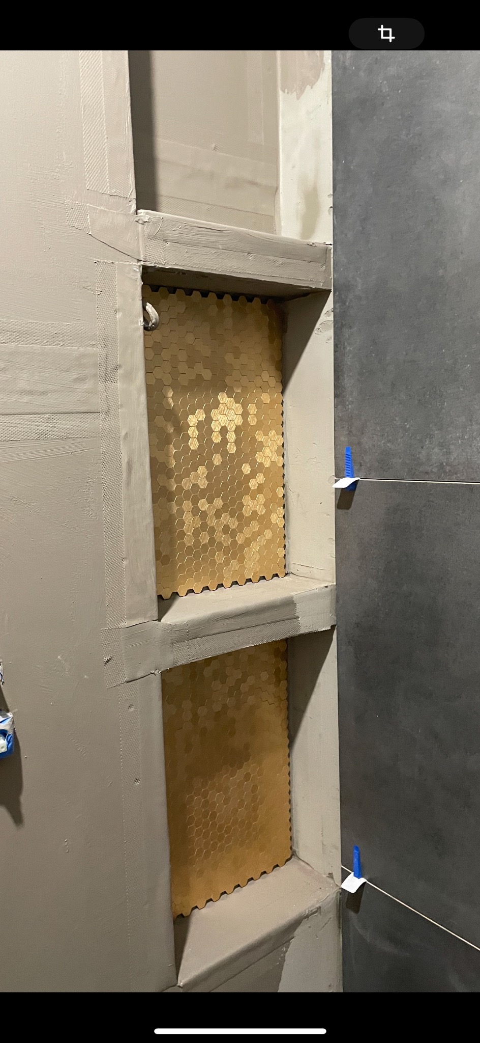 Półki wnękowe w łazience wyłożone mozaiką w kształcie złotych heksagonów, obok ściana z ciemnoszarych płytek z poziomą fugą, zabezpieczona niebieskim klinem dystansowym.