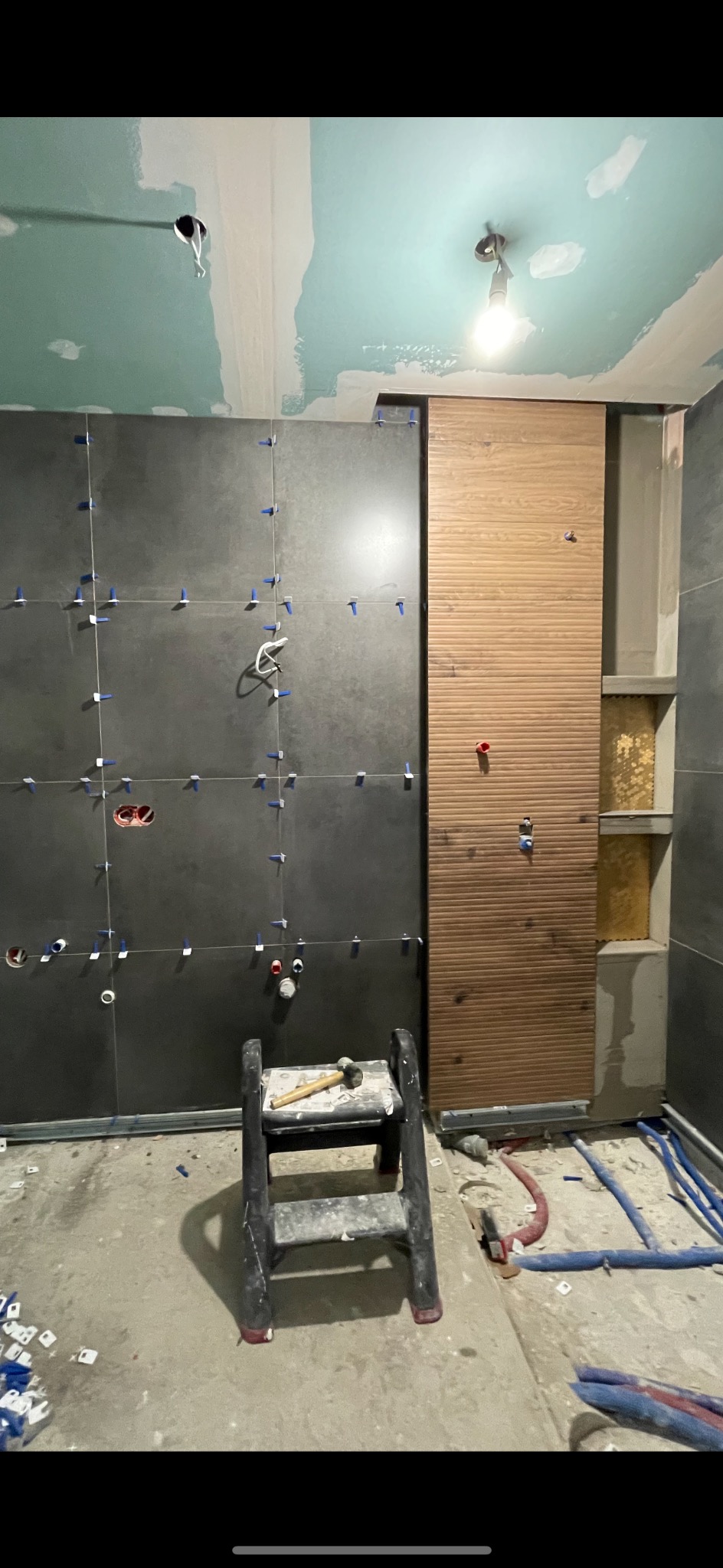 Ściana łazienki w trakcie układania płytek ceramicznych w odcieniach szarości i drewnopodobnych, widoczne krzyżyki dystansowe, drabinka i instalacje hydrauliczne.