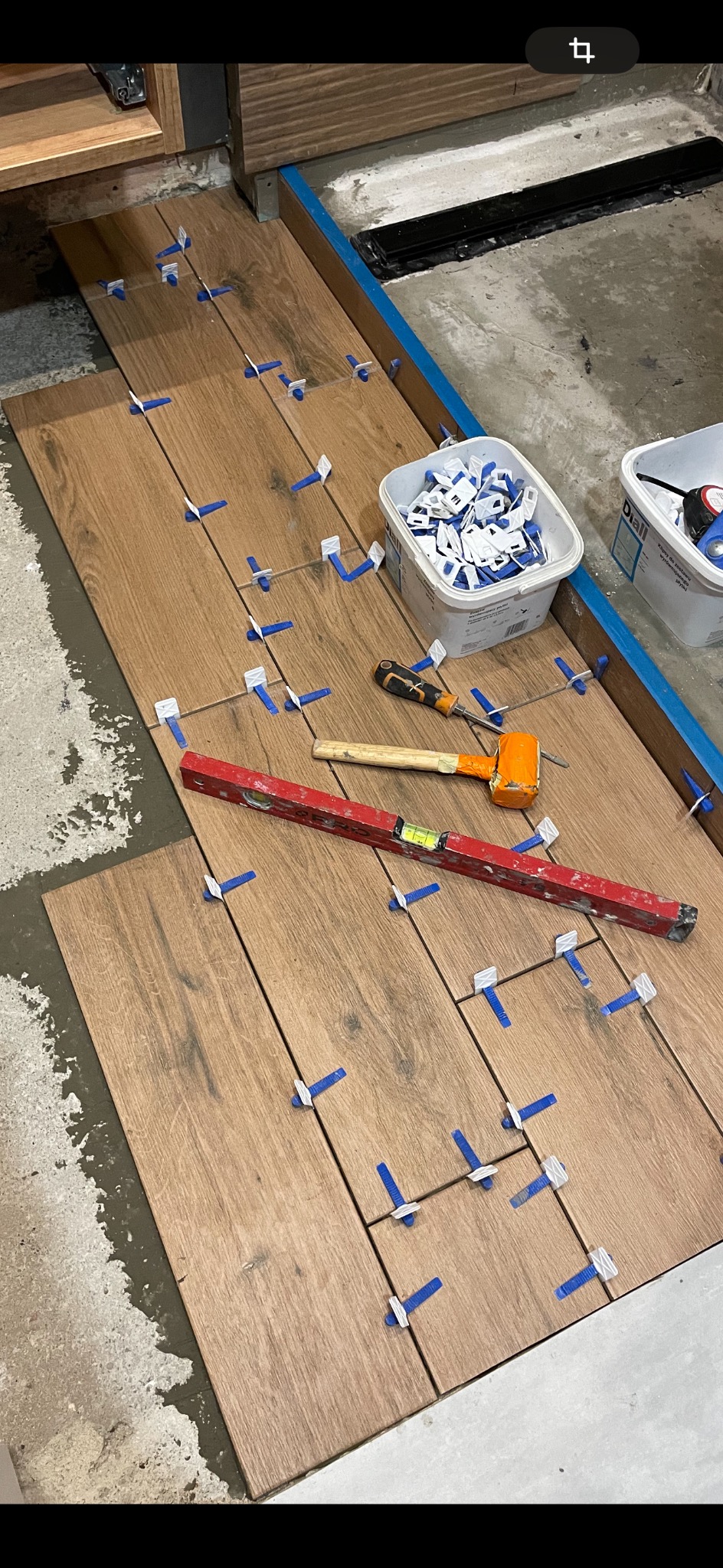 Układanie płytek imitujących drewno na podłodze, widoczne poziomica, gumowy młotek i plastikowe krzyżyki dystansowe, na podłodze resztki zaprawy