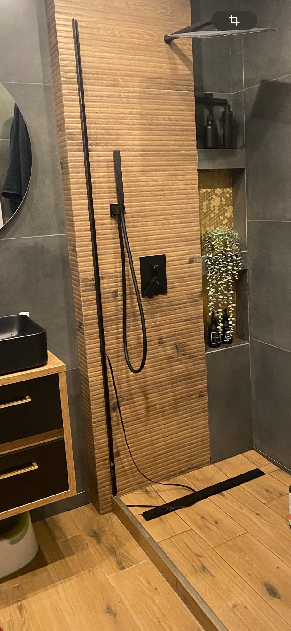 Nowoczesna kabina prysznicowa z drewnopodobnymi płytkami na ścianie i podłodze, czarna armatura i panel sterujący, dekoracyjna półka z rośliną zwisającą i kosmetykami.