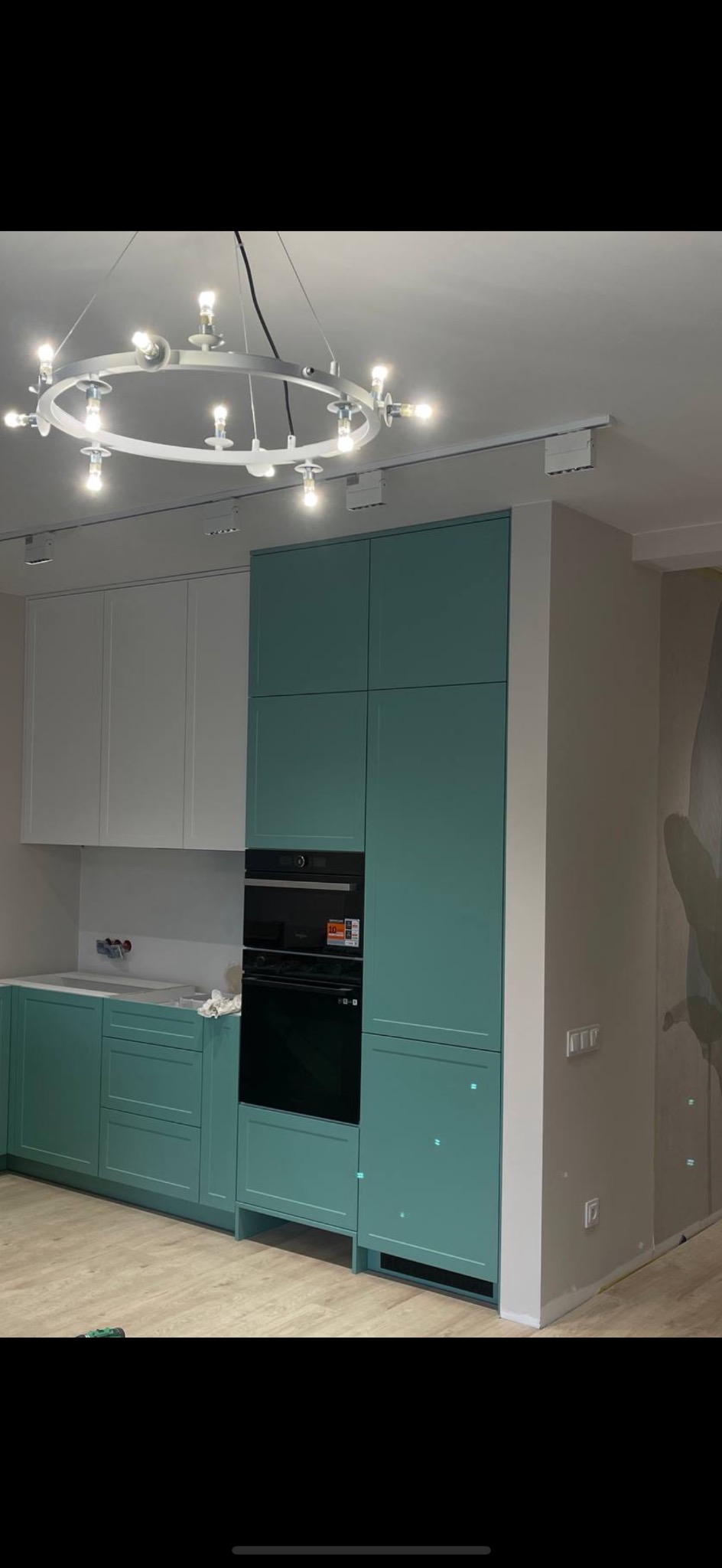 Nowoczesna kuchnia z turkusowymi szafkami dolnymi i wysokimi, białymi szafkami górnymi, czarnym piekarnikiem w zabudowie, okrągłym żyrandolem i szynoprzewodem sufitowym, widoczne ślady prac...
