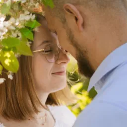 Zbliżenie na profil kobiety w okularach i słomkowym kapeluszu, otoczonej kwitnącymi gałązkami, dotykającej czołem mężczyzny z brodą w błękitnej koszuli.