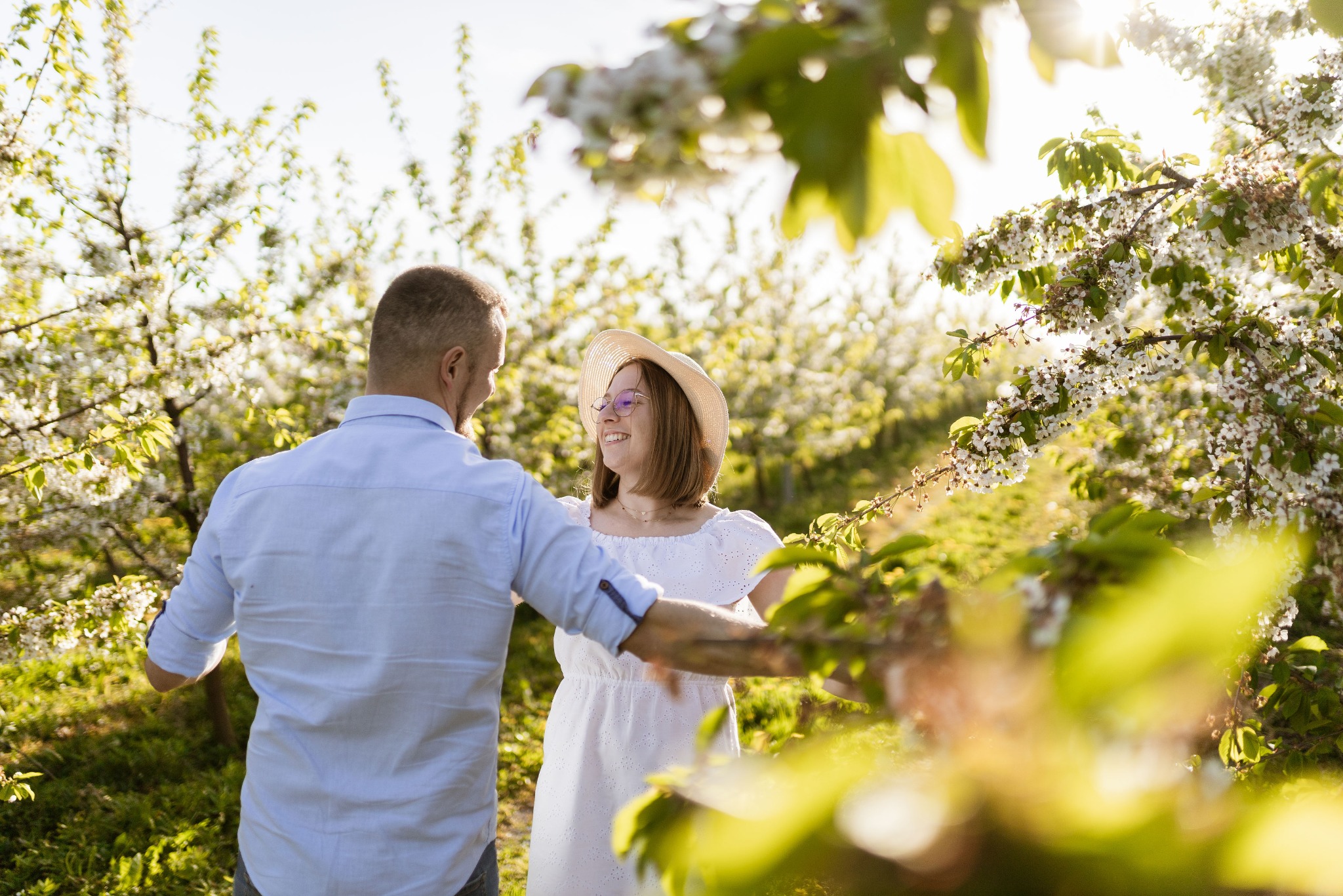 Para w kwitnącym sadzie; mężczyzna w niebieskiej koszuli obejmuje uśmiechniętą kobietę w białej sukience i kapeluszu, oboje patrzą na siebie w słoneczny dzień.