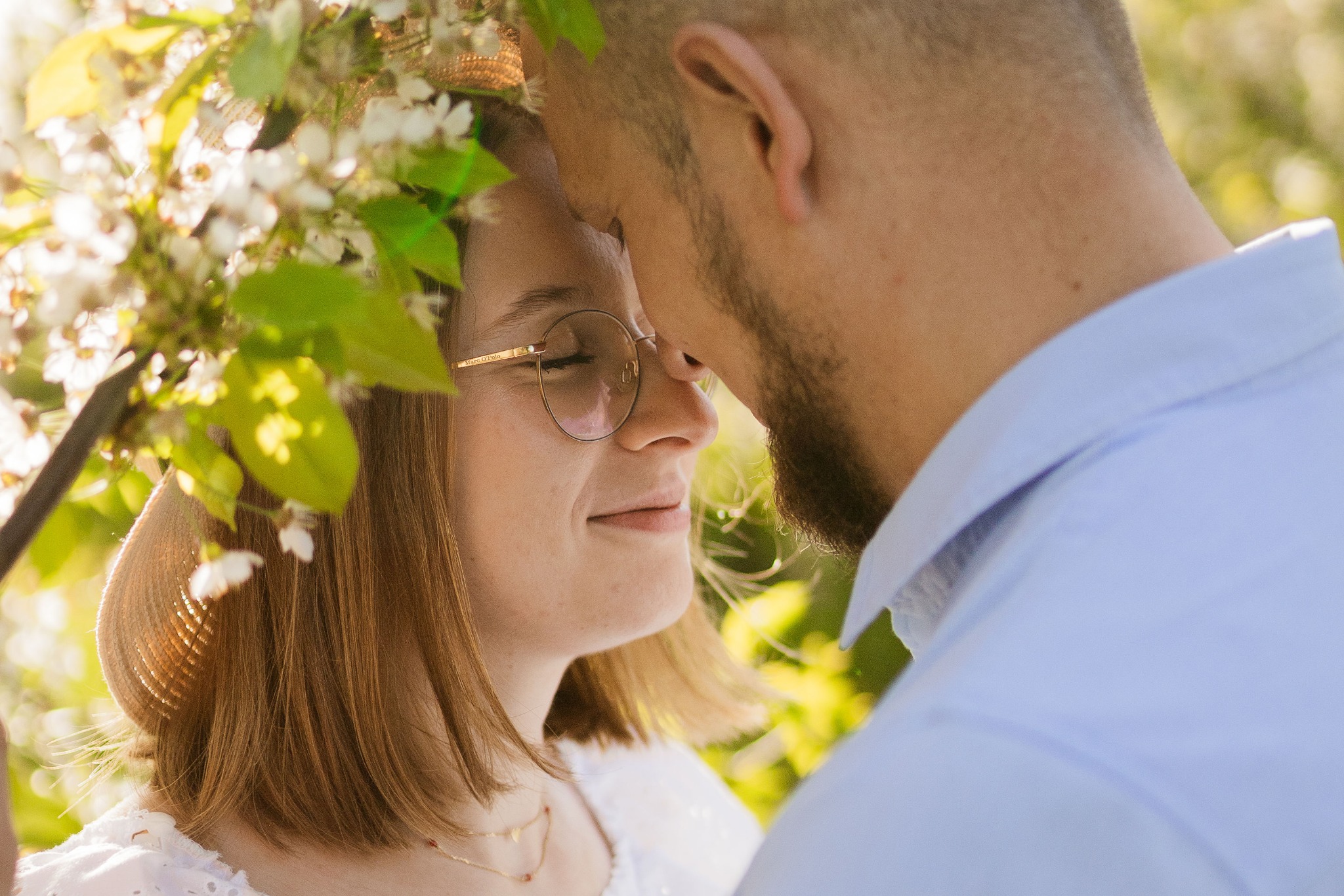 Zbliżenie na profil kobiety w okularach i słomkowym kapeluszu, otoczonej kwitnącymi gałązkami, dotykającej czołem mężczyzny z brodą w błękitnej koszuli.