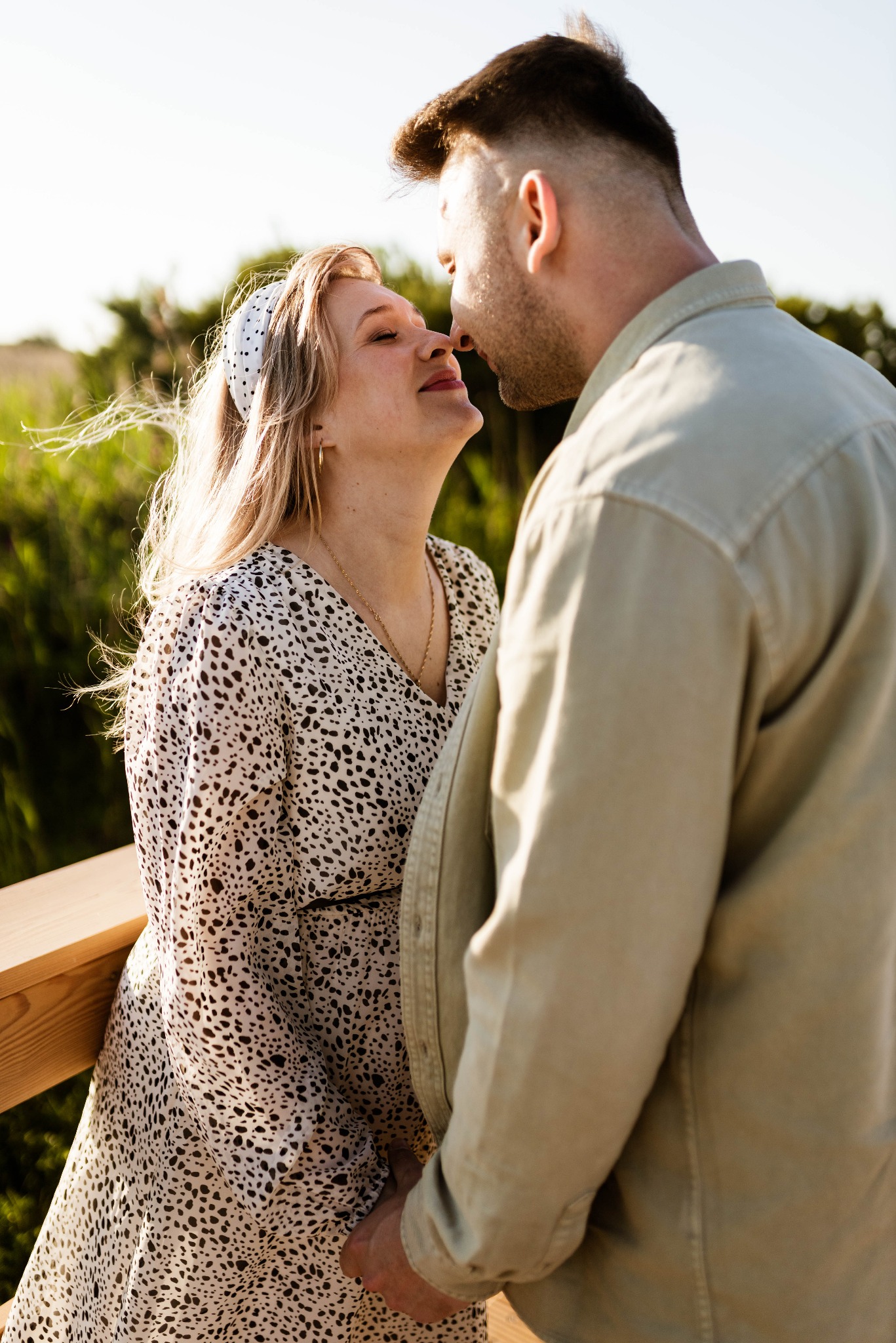 Para zakochanych, kobieta w sukience w panterkę i opasce we włosach, mężczyzna w beżowej koszuli, zbliżają się do pocałunku na drewnianym pomoście, zielone tło roślinności.
