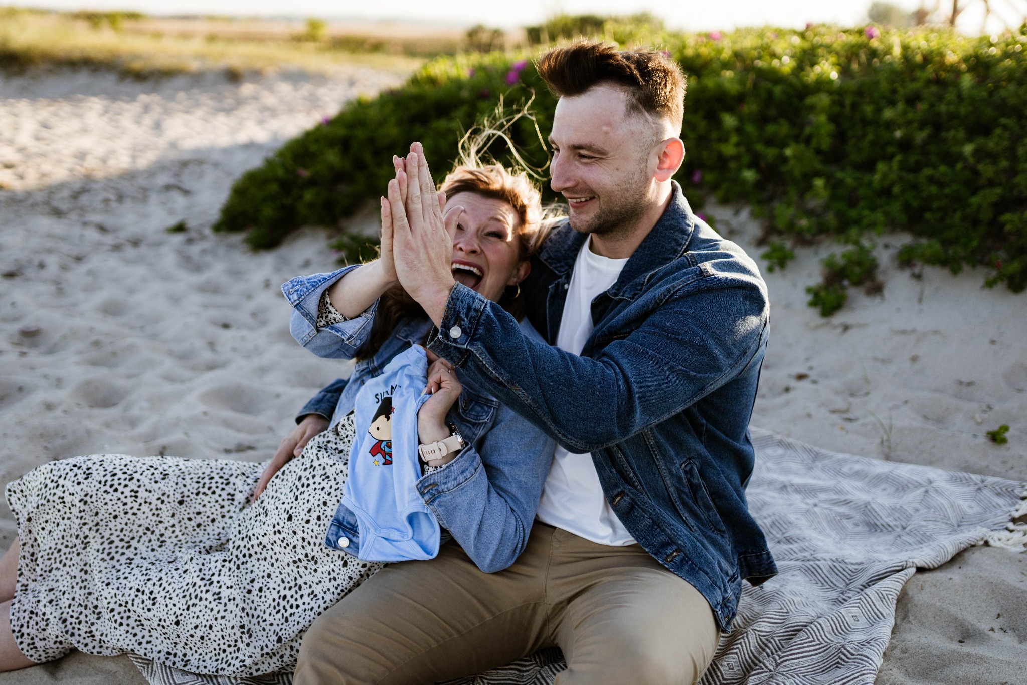 Uśmiechnięta para siedzi na plaży na kocu, kobieta w kwiecistej sukience trzyma niebieskie niemowlęce body z nadrukiem Supermana, mężczyzna w jeansowej kurtce przybija z nią piątkę, w tle piasek...