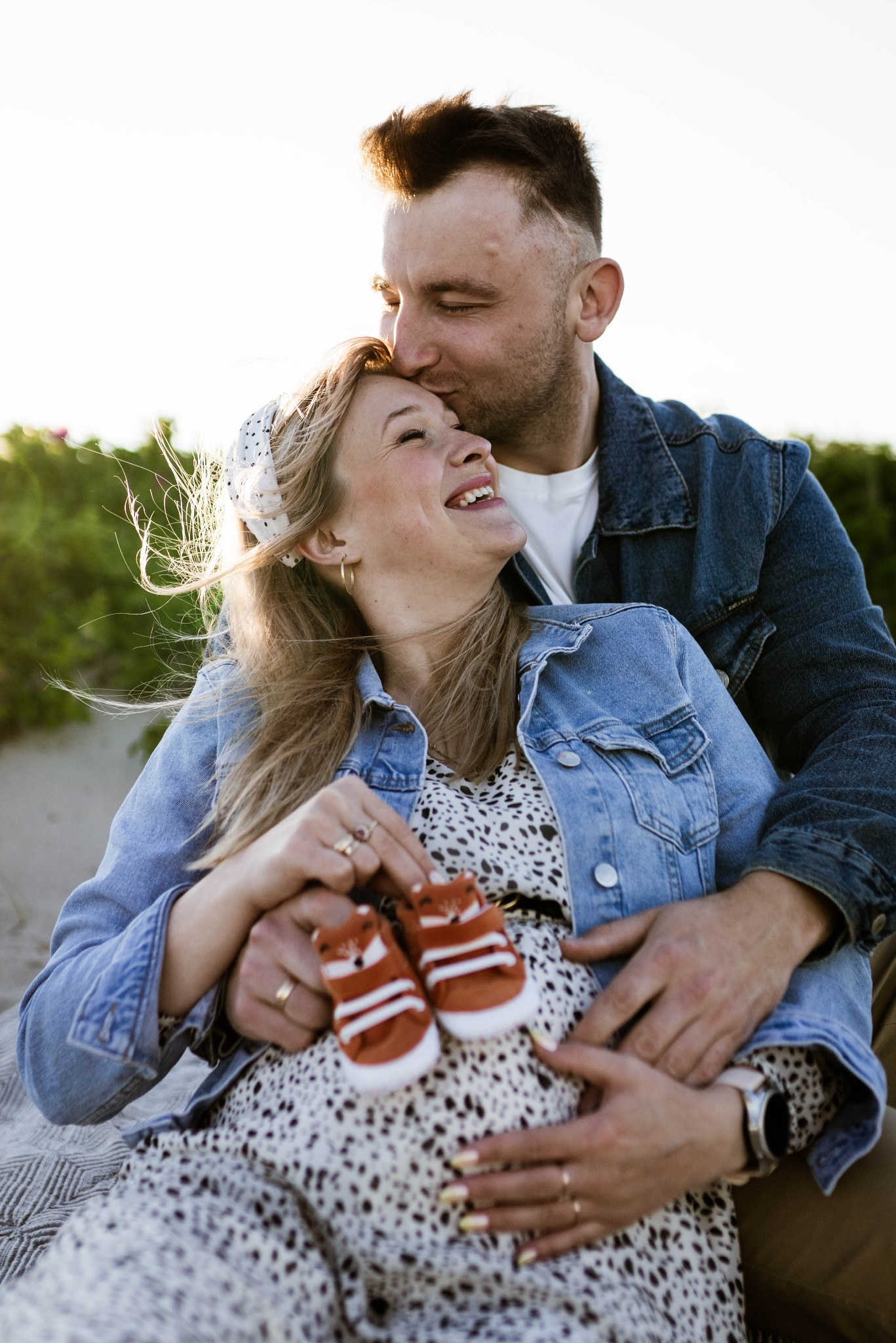 Uśmiechnięta, ciężarna kobieta w sukience w panterkę i dżinsowej kurtce, siedząca na kocu, trzymająca małe, lisie buciki na brzuchu, obejmowana przez mężczyznę w dżinsowej kurtce, który całuje ją...