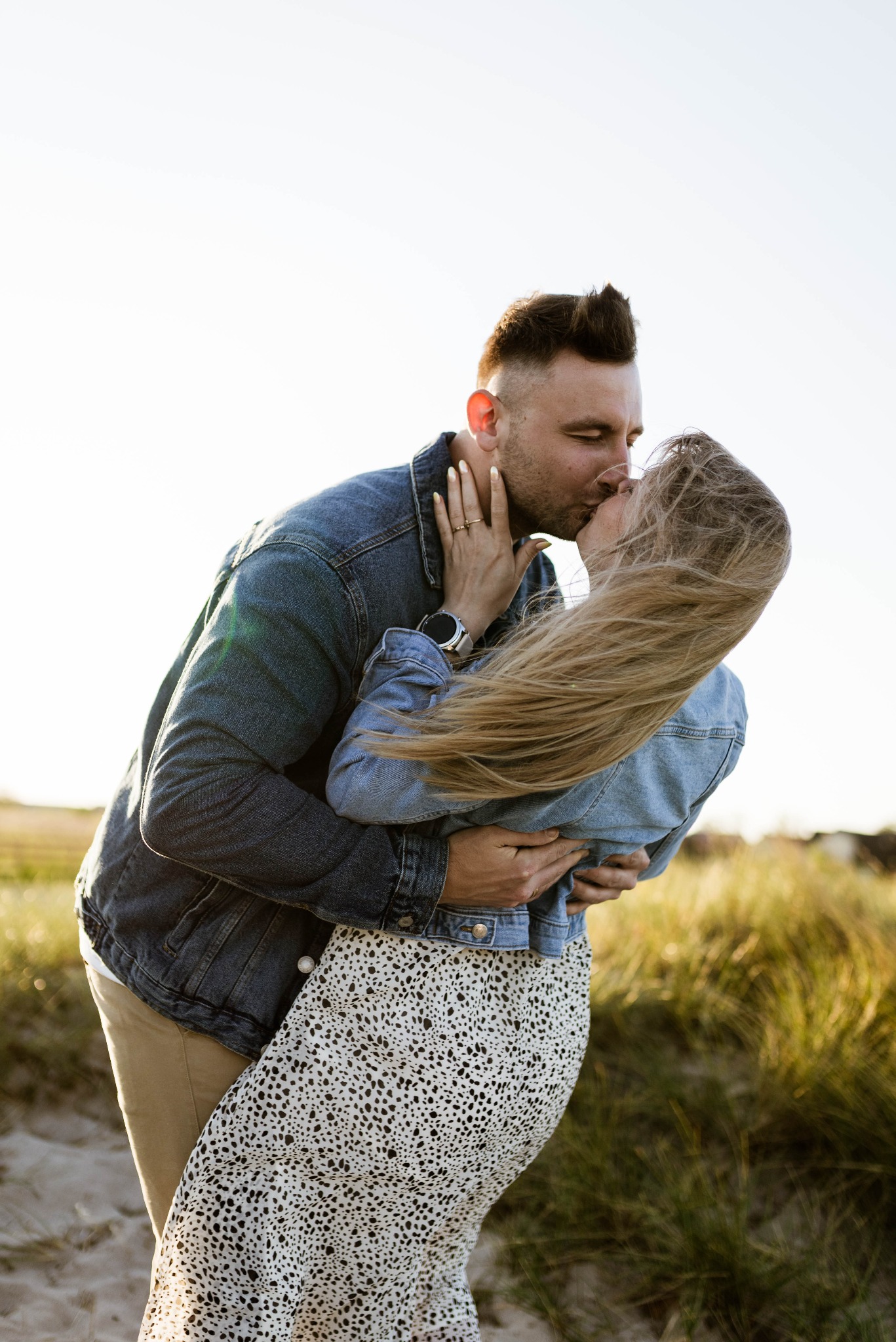 Para całująca się na tle trawy i jasnego nieba, kobieta w kwiecistej sukience i jeansowej kurtce, mężczyzna w jeansowej kurtce i beżowych spodniach, widoczny zegarek na ręce kobiety.