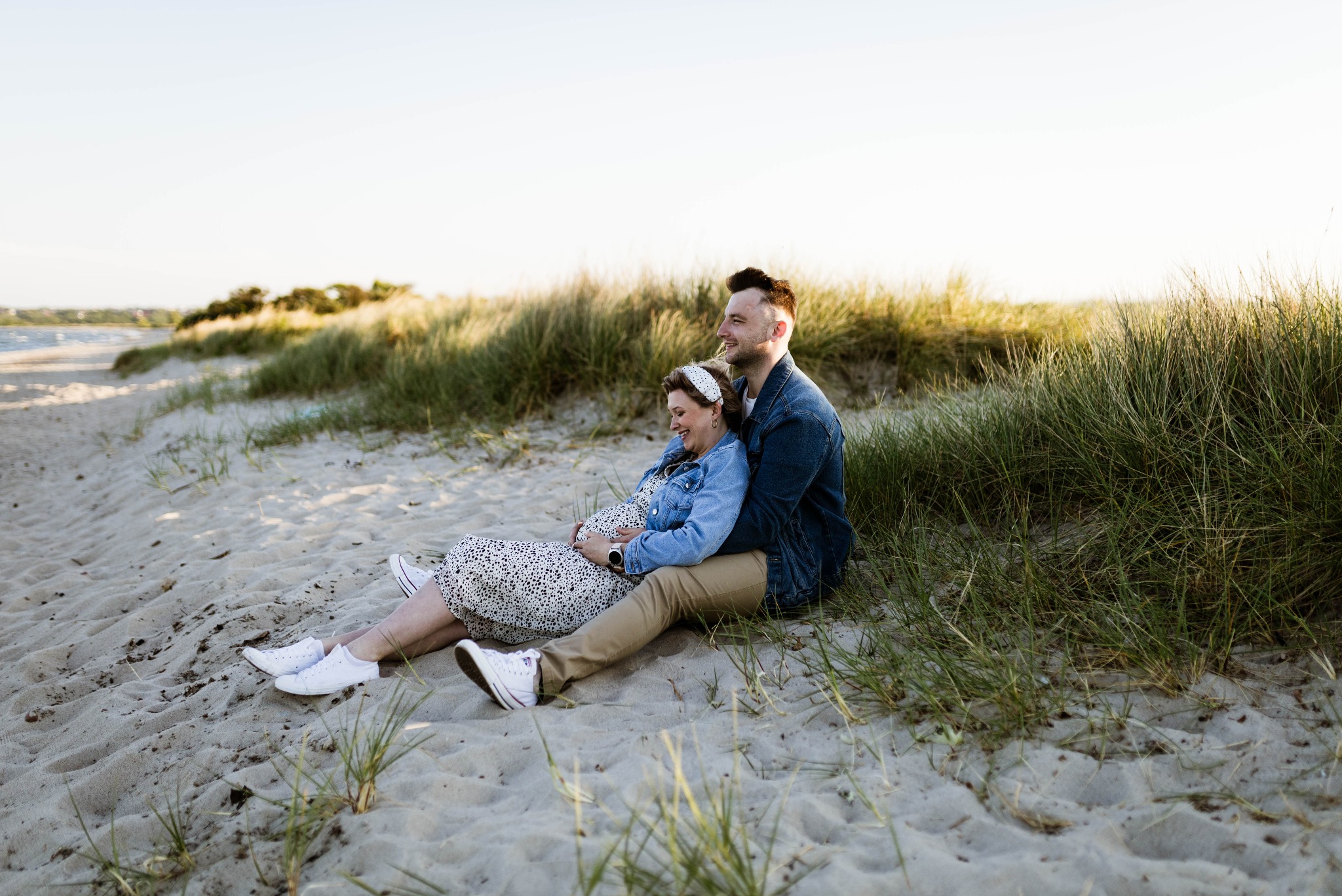 Sesja narzeczeńska na plaży: uśmiechnięta para siedzi na piasku, kobieta w kwiecistej sukience i dżinsowej kurtce trzyma się za ciążowy brzuszek, mężczyzna w dżinsowej kurtce obejmuje ją, w tle...