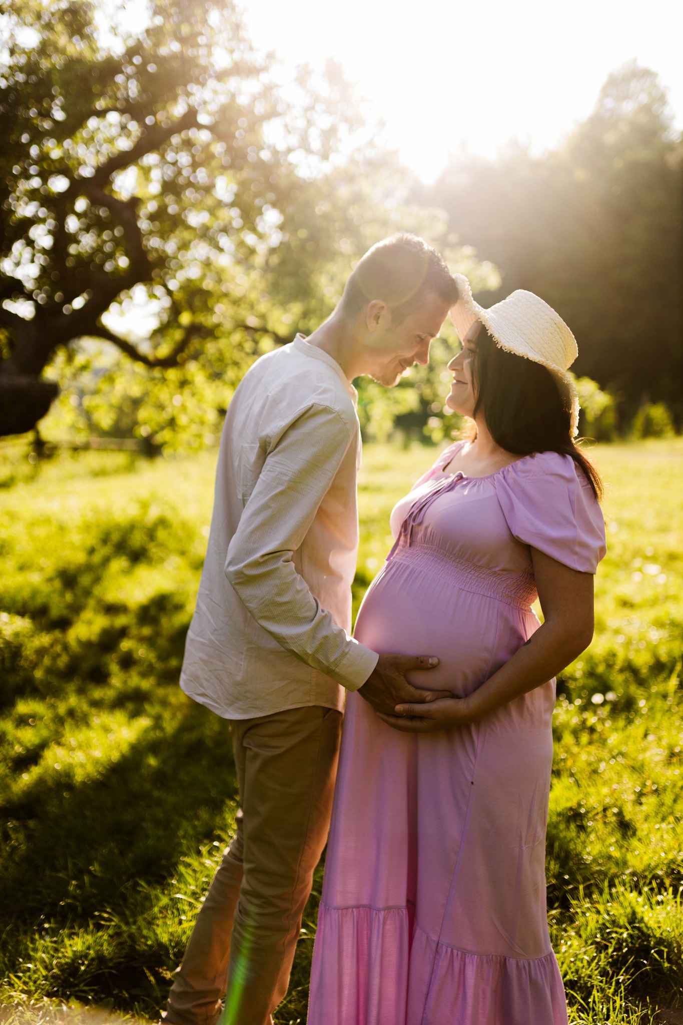 Przyszła mama w długiej, liliowej sukience i słomkowym kapeluszu, obejmowana przez mężczyznę na tle zieleni parkowej w promieniach zachodzącego słońca.