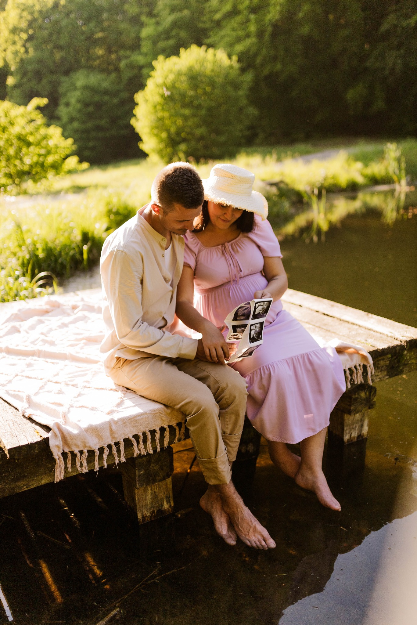 Przyszli rodzice siedzą na drewnianym pomoście nad wodą, oglądają zdjęcia USG dziecka, kobieta w liliowej sukience i kapeluszu, mężczyzna w beżowej koszuli i spodniach.
