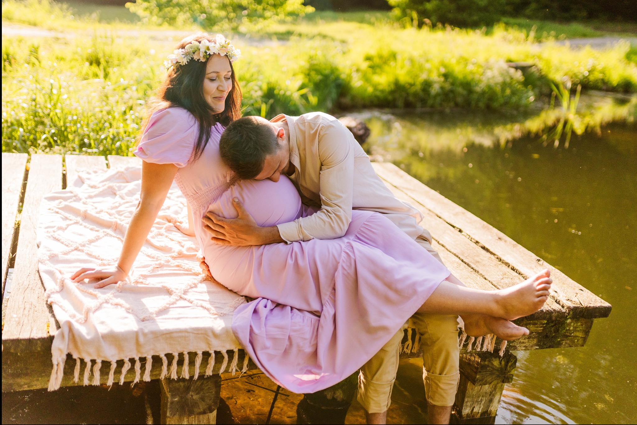 Kobieta w kwiecistym wianku i liliowej sukience siedzi na pomoście nad wodą, mężczyzna w beżowej koszuli obejmuje jej brzuch.