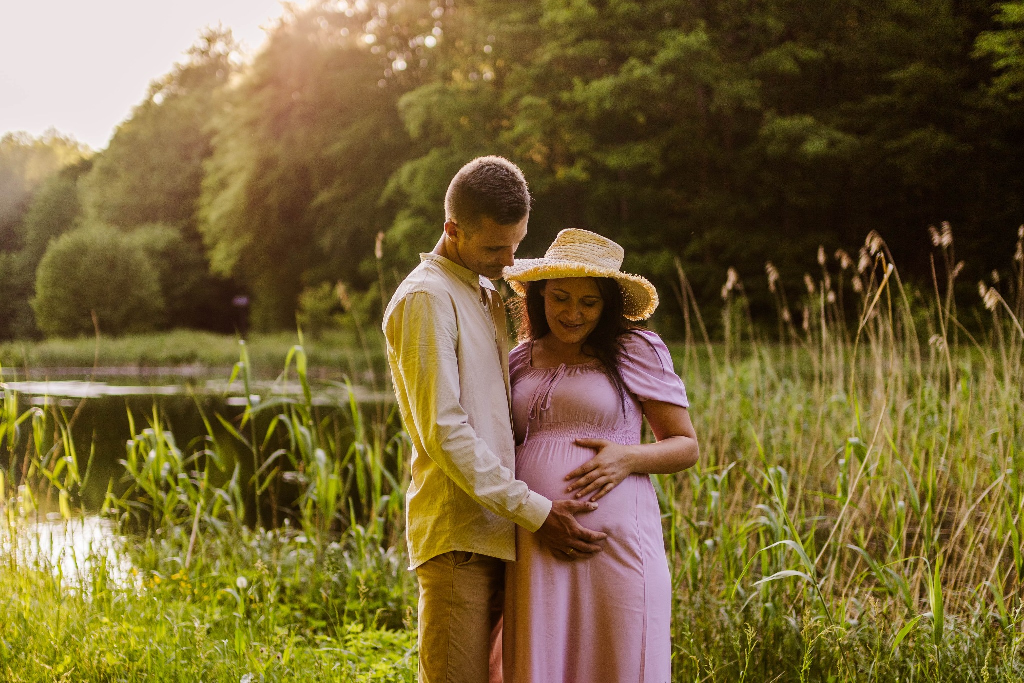 Przyszła mama w długiej, liliowej sukience i słomkowym kapeluszu, obejmowana przez partnera na tle jeziora i zieleni, podczas sesji ciążowej w plenerze.