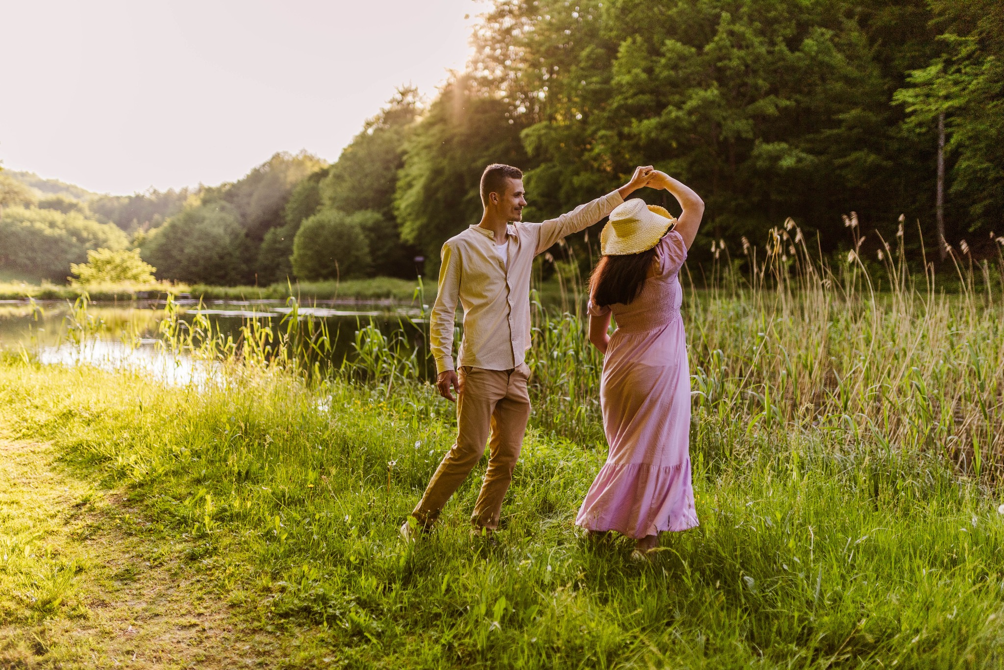 Para tańczy na łące w pobliżu jeziora o zachodzie słońca. Mężczyzna w beżowej koszuli i spodniach trzyma kobietę w długiej, różowej sukience i słomkowym kapeluszu.