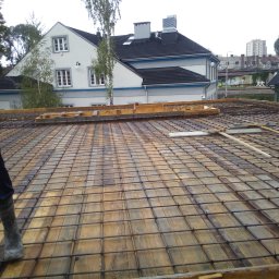 ZRBH Dragon Paweł - Zbrojenie stropu z siatki stalowej na deskowaniu drewnianym, widoczny fragment sylwetki robotnika w roboczych butach i rękawicy, w tle budynek mieszkalny.