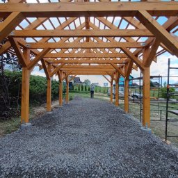 ZRBH Dragon Paweł - Drewniana konstrukcja z belek i słupów na betonowych fundamentach, widok z wnętrza na zewnątrz, osoba w tle, teren wysypany żwirem.