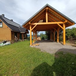 ZRBH Dragon Paweł - Drewniana wiata garażowa z szarym dachem, widoczna z zewnątrz na tle domu z panelami słonecznymi i zieleni, na żwirowym podłożu, z ułożonym drewnem w tle.