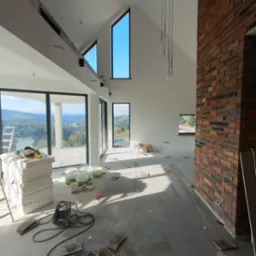 Wnętrze domu w trakcie wykańczania: murowana ściana z cegły, duże okna z widokiem na krajobraz, rozłożone narzędzia i materiały budowlane na podłodze.