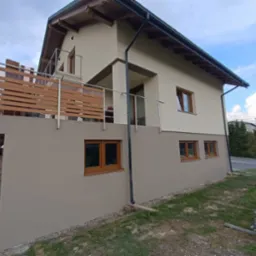 Dwukondygnacyjny dom z beżową elewacją i drewnianymi oknami, balkon z drewnianą balustradą, widoczne rusztowania i teren w trakcie zagospodarowania.