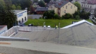 Prace na dachu płaskim budynku z widocznymi dwoma pracownikami i materiałami budowlanymi, w tle zabudowa miejska i zieleń.