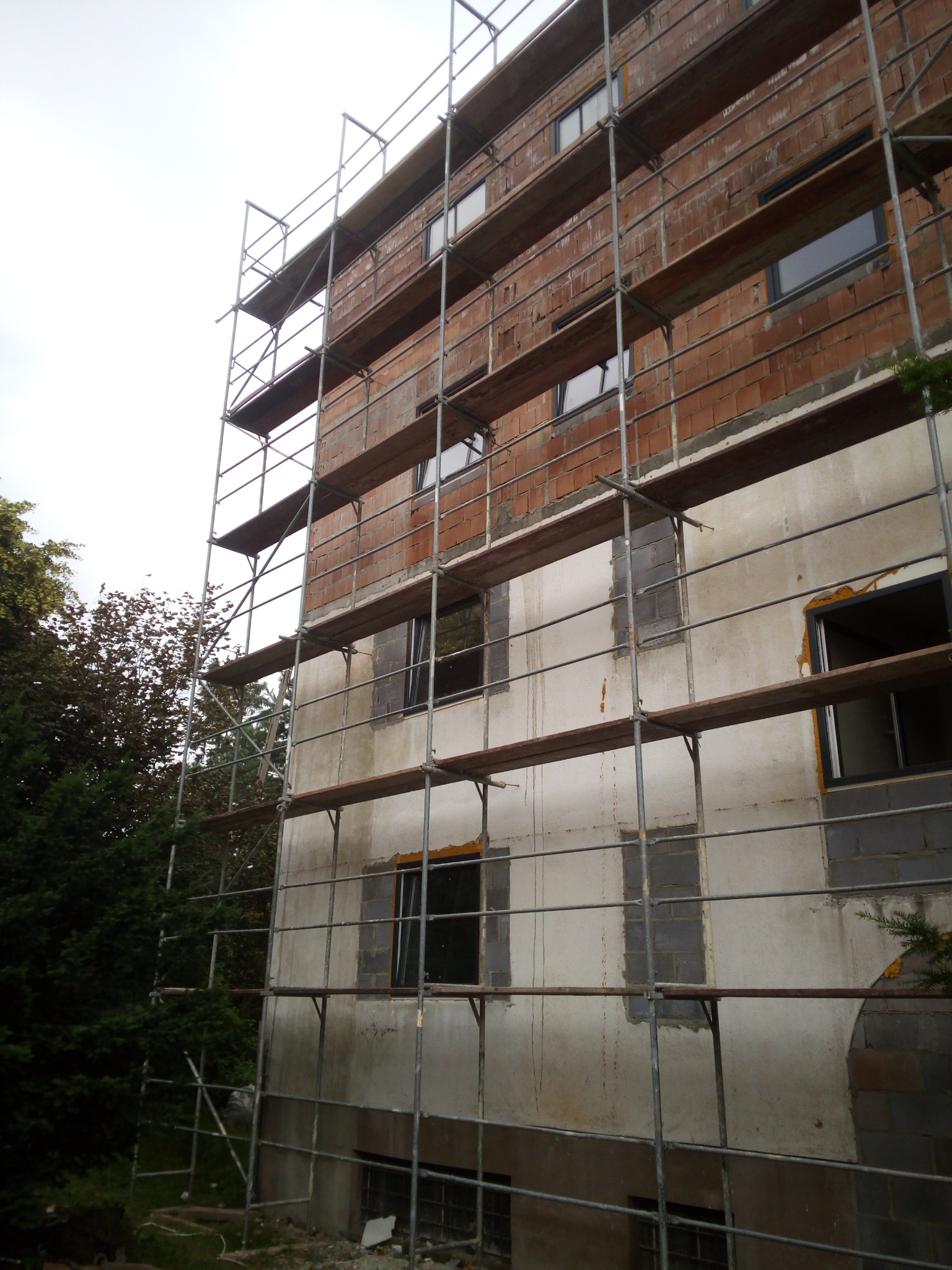Budynek w trakcie prac budowlanych, widoczne rusztowania, fragmenty murowanej ściany z cegły oraz otynkowanej elewacji z przygotowanymi otworami okiennymi.