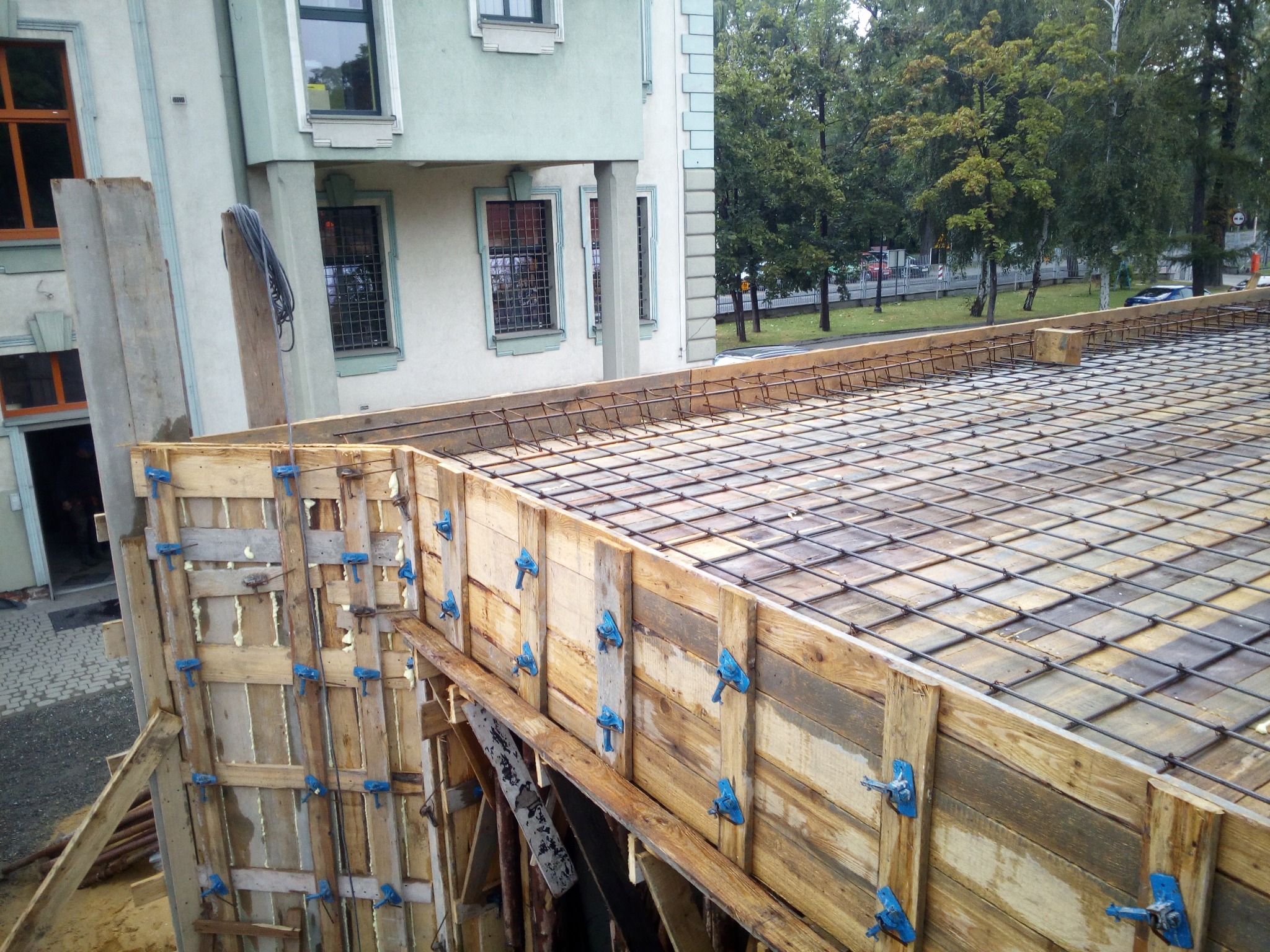 Szczegółowy widok szalunku zbrojeniowego na budowie domu, z drewnianymi deskami i niebieskimi zaciskami, przygotowanego do wylania betonu, na tle zieleni drzew i fragmentu budynku.