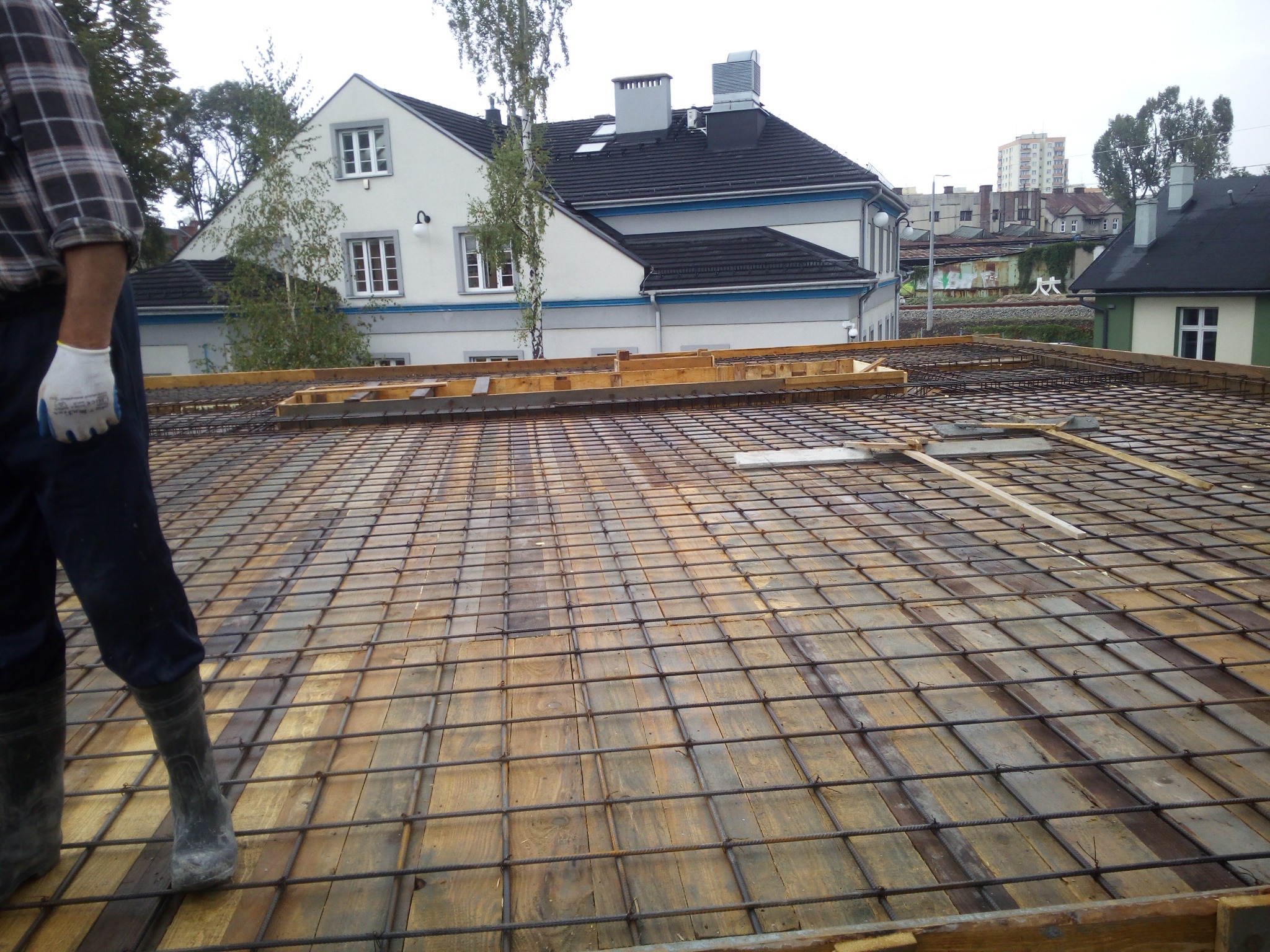 Zbrojenie stropu z siatki stalowej na deskowaniu drewnianym, widoczny fragment sylwetki robotnika w roboczych butach i rękawicy, w tle budynek mieszkalny.