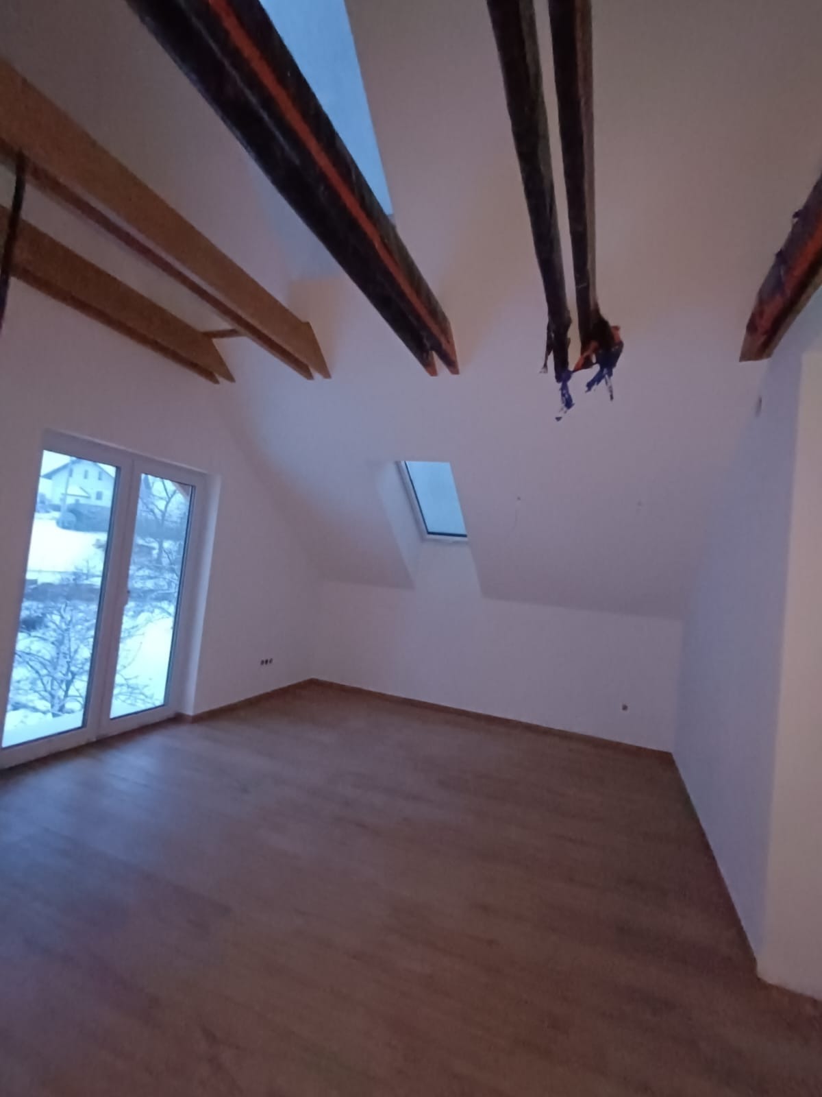 Poddasze w Jaworzynce po remoncie: drewniane belki stropowe, okno dachowe i duże okno szczytowe z widokiem na zimowy krajobraz, świeżo położona podłoga.