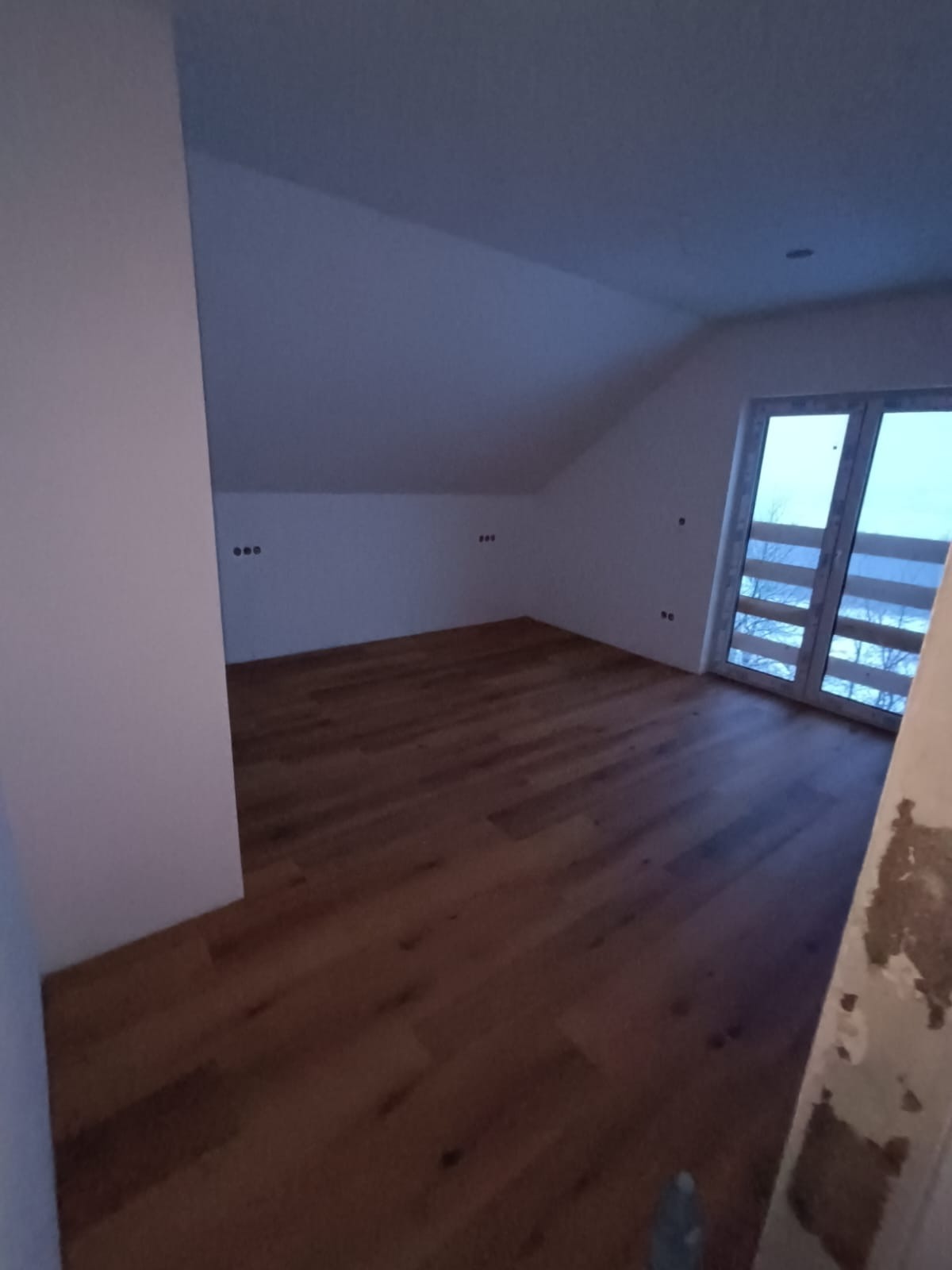 Poddasze z drewnianą podłogą w jodełkę, świeżo pomalowane ściany na biało, widoczne gniazdka elektryczne i okno balkonowe z widokiem na zewnątrz.