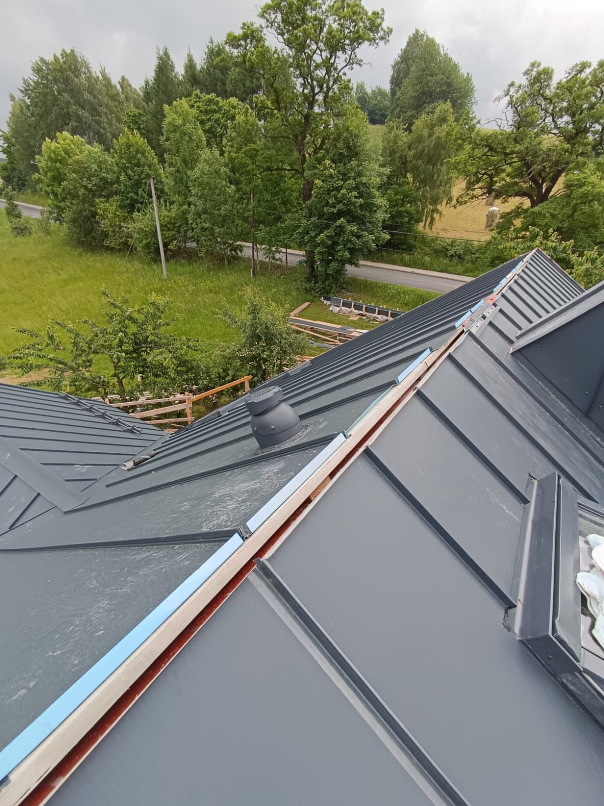 Szary blaszany dach domu jednorodzinnego z widoczną wentylacją kominową i elementami montażowymi, na tle zieleni i drogi.