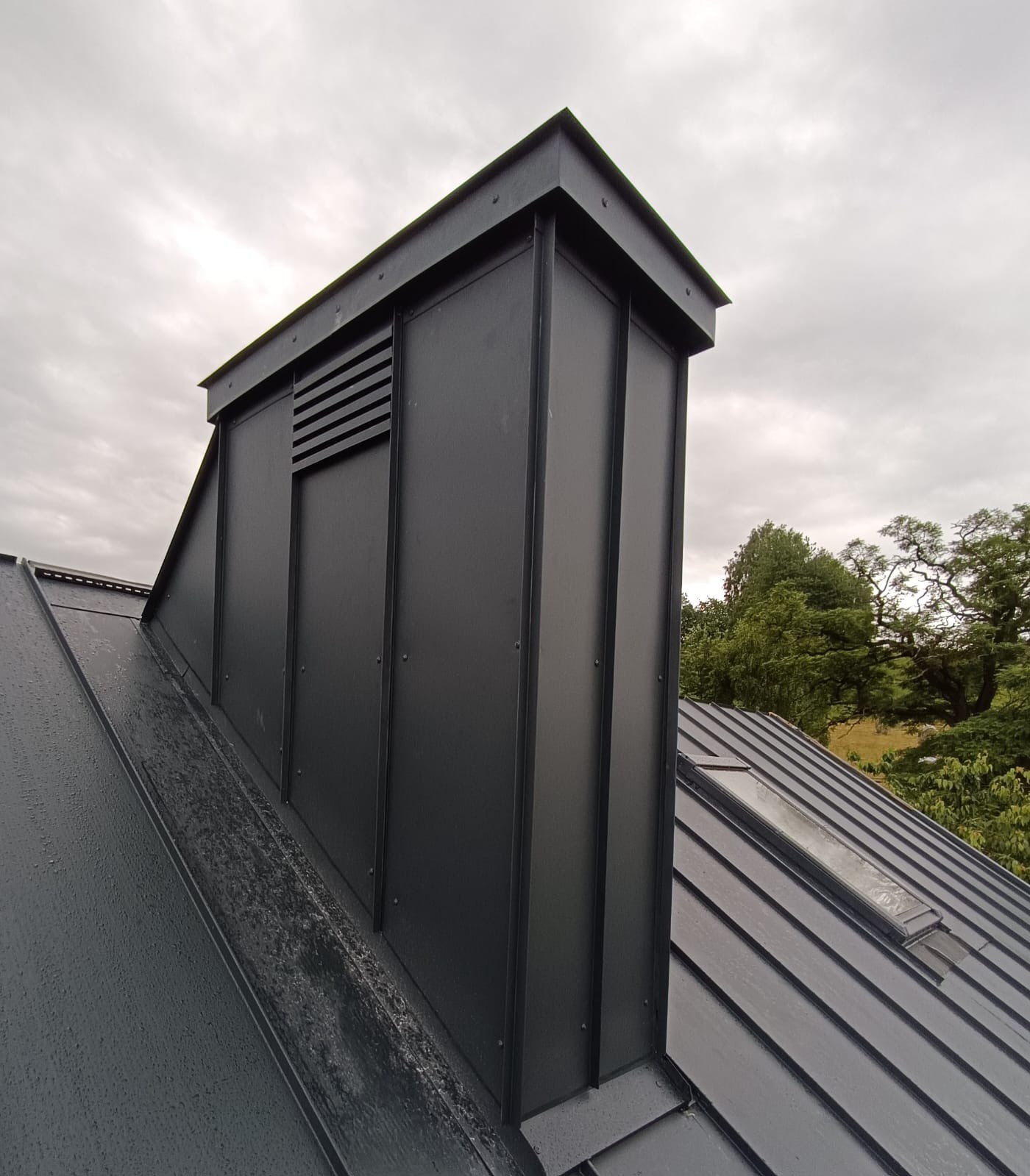 Nowoczesny, czarny komin wentylacyjny z poziomymi kratkami na metalowym dachu z oknem dachowym, widok z dołu na tle pochmurnego nieba i zieleni drzew.