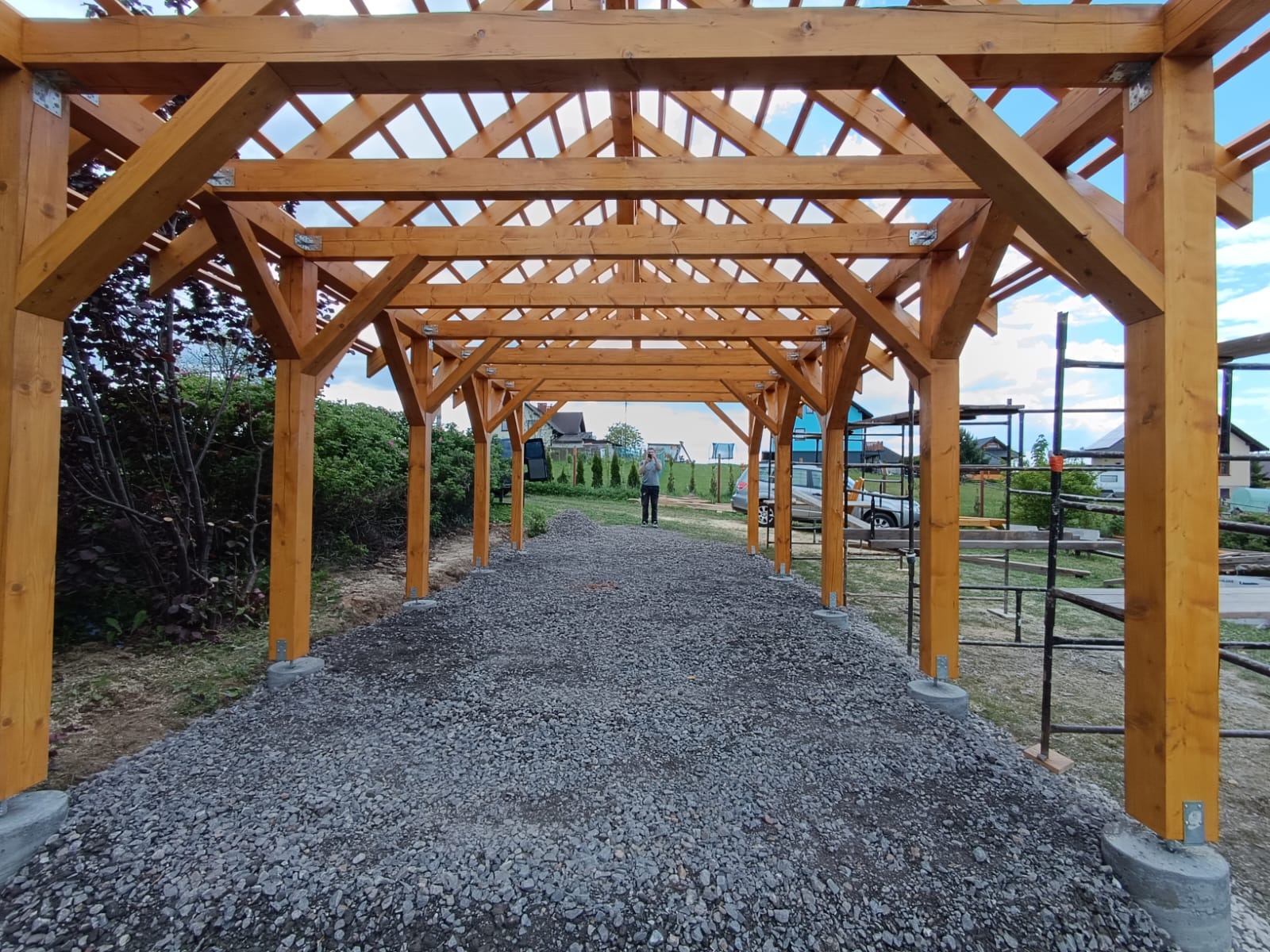 Drewniana konstrukcja z belek i słupów na betonowych fundamentach, widok z wnętrza na zewnątrz, osoba w tle, teren wysypany żwirem.