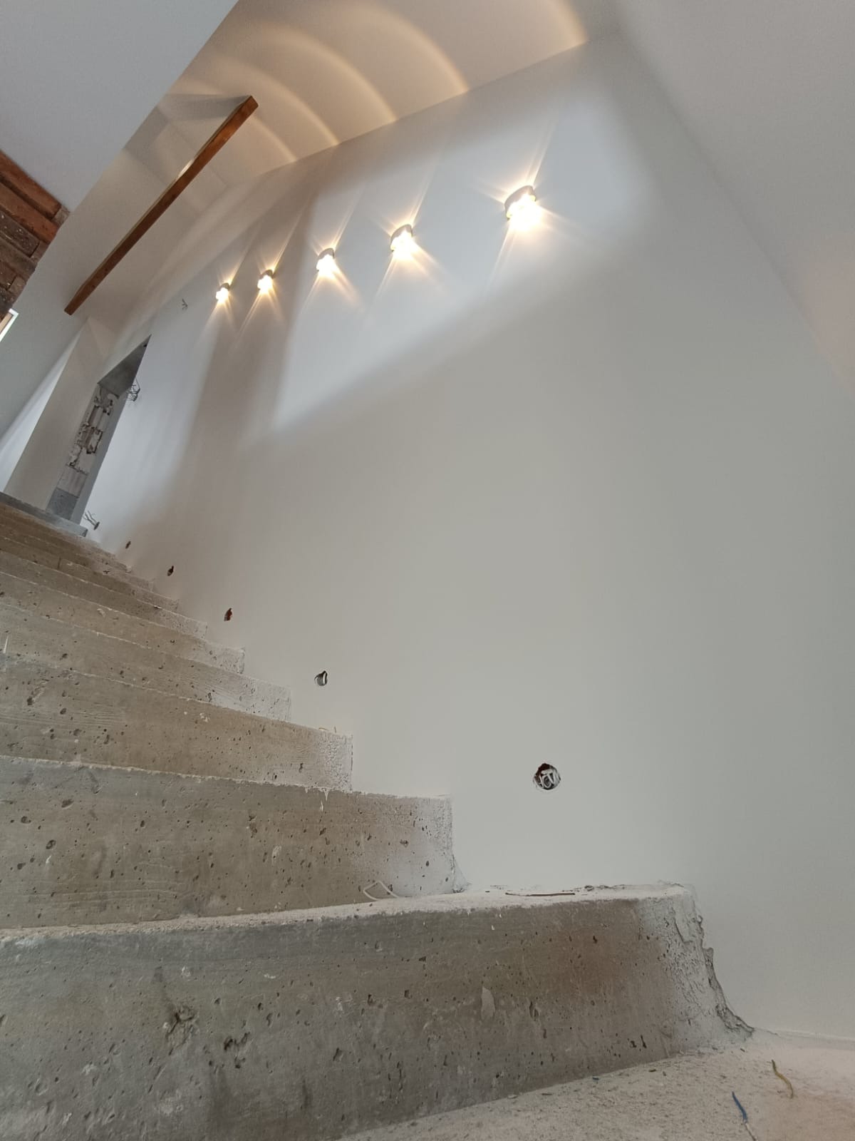 Surowe betonowe schody wewnętrzne w trakcie budowy, widoczne puszki elektryczne w ścianie i rząd lamp sufitowych.