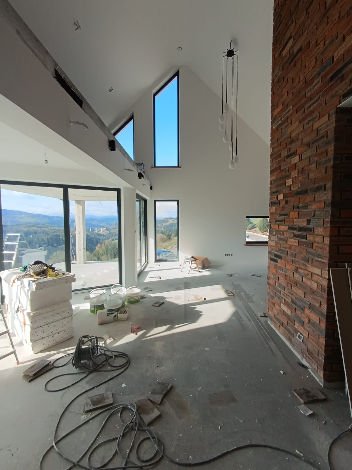 Wnętrze domu w trakcie wykańczania: murowana ściana z cegły, duże okna z widokiem na krajobraz, rozłożone narzędzia i materiały budowlane na podłodze.