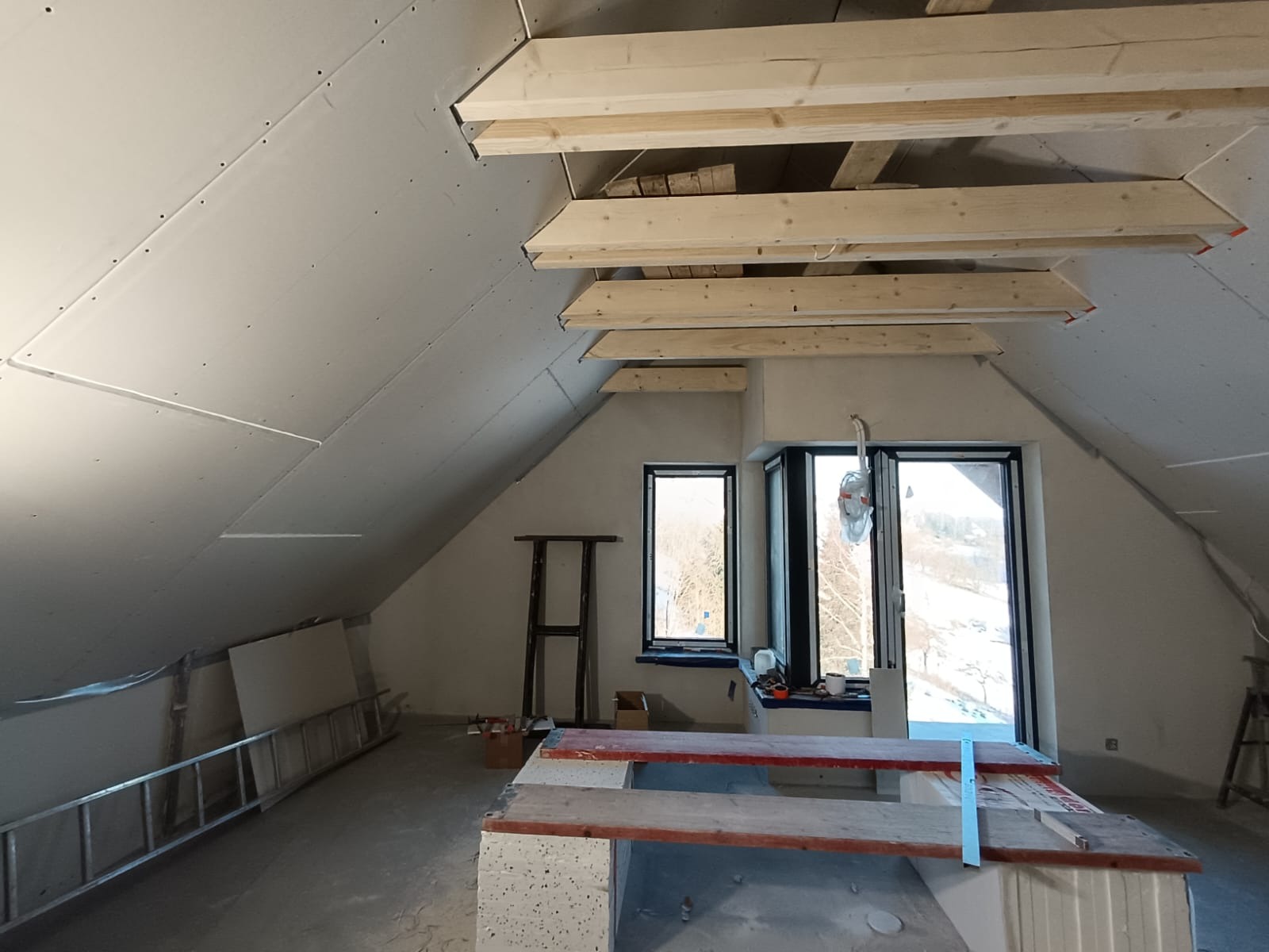 Poddasze w trakcie wykańczania: widoczne płyty gipsowo-kartonowe na skośnym suficie, drewniane belki konstrukcyjne, okna z widokiem na zewnątrz, drabina oparta o ścianę, stanowisko robocze...