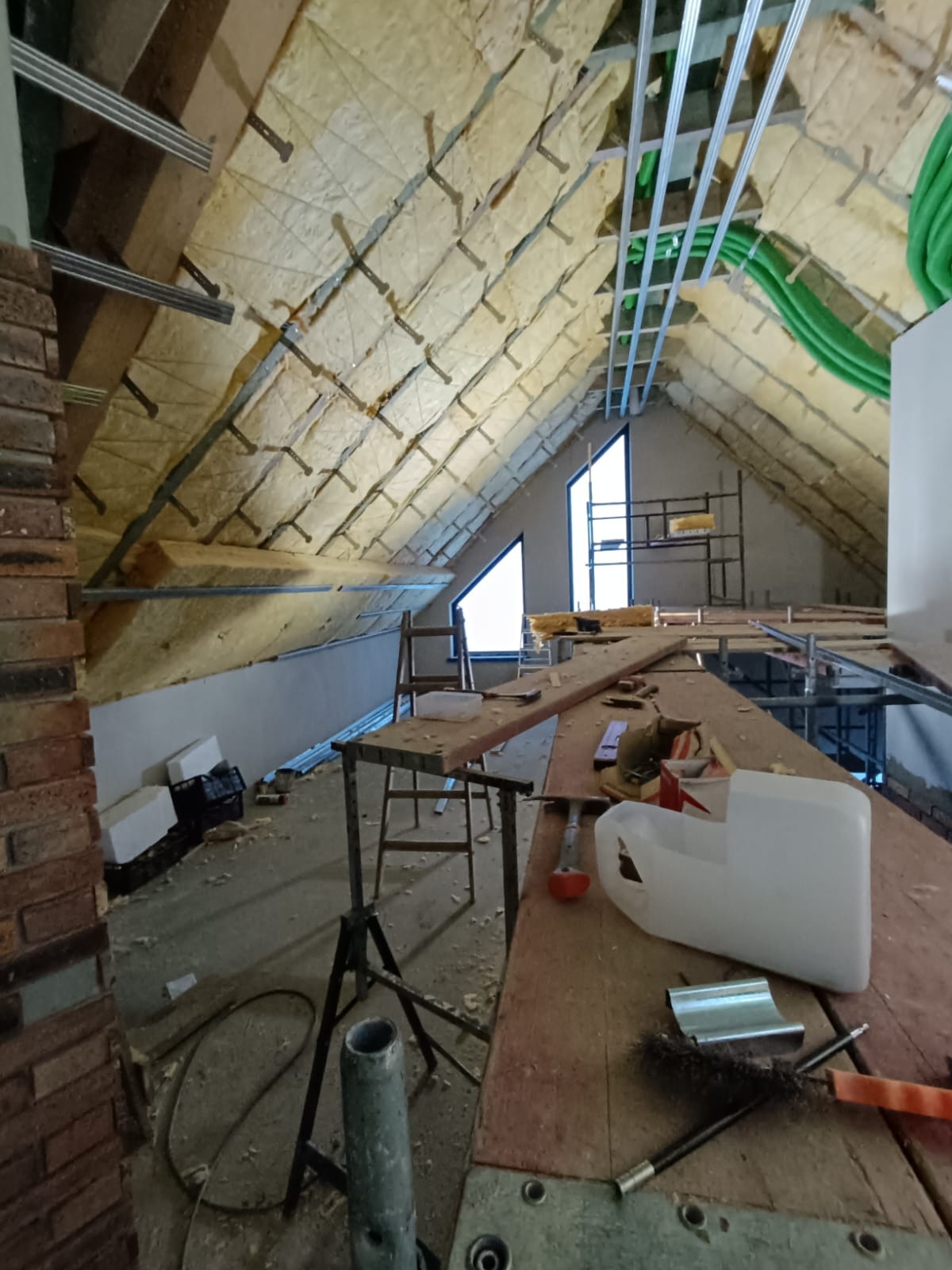 Poddasze w trakcie ocieplania: widoczna wełna mineralna między krokwiami, metalowe profile pod płyty gipsowe, fragment instalacji wentylacyjnej, okna dachowe i prowizoryczny stół roboczy...