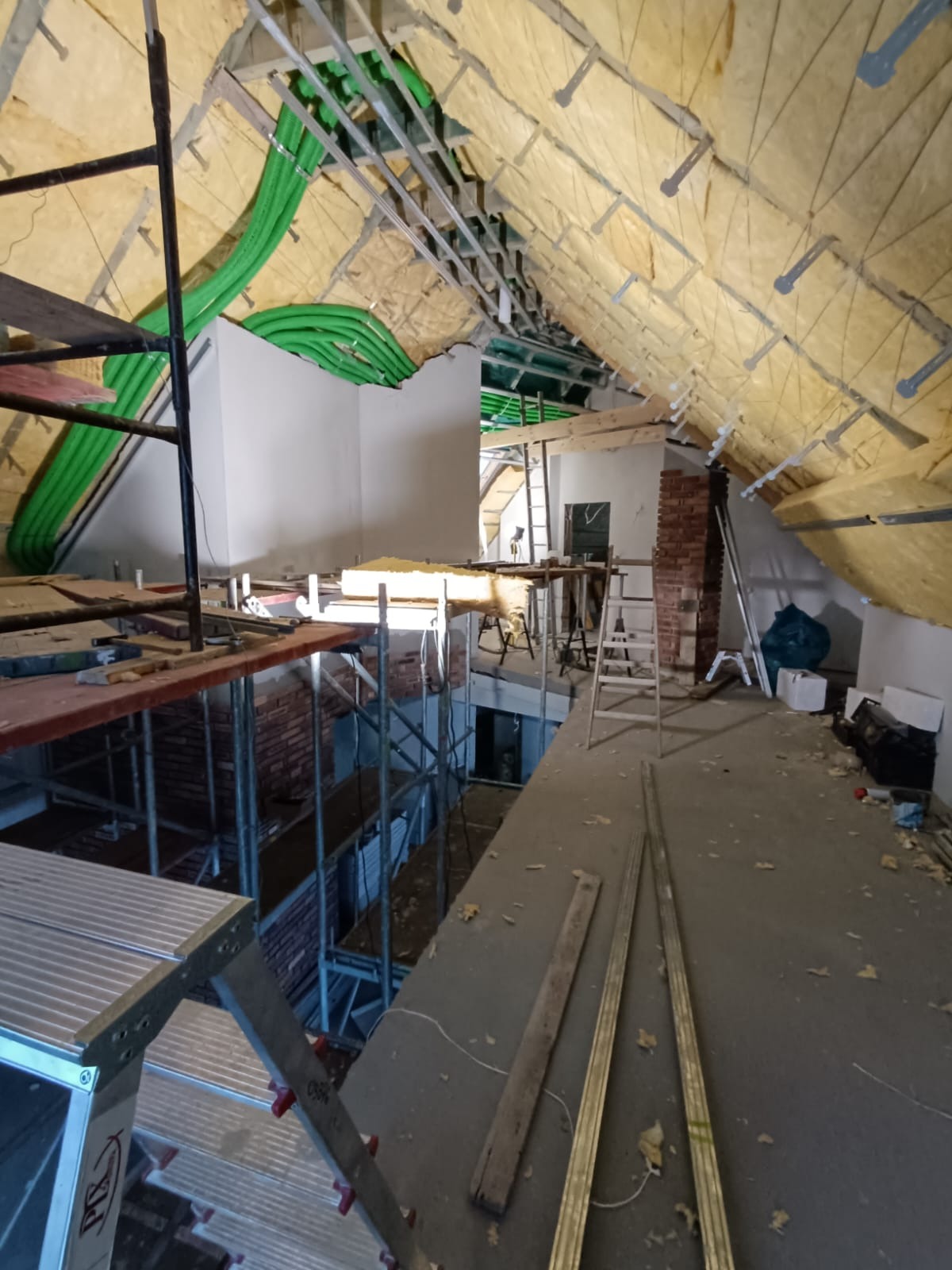 Poddasze w trakcie adaptacji: widoczne rusztowania, fragment ściany z cegły, izolacja stropu wełną mineralną oraz instalacja wentylacyjna z zielonych rur.